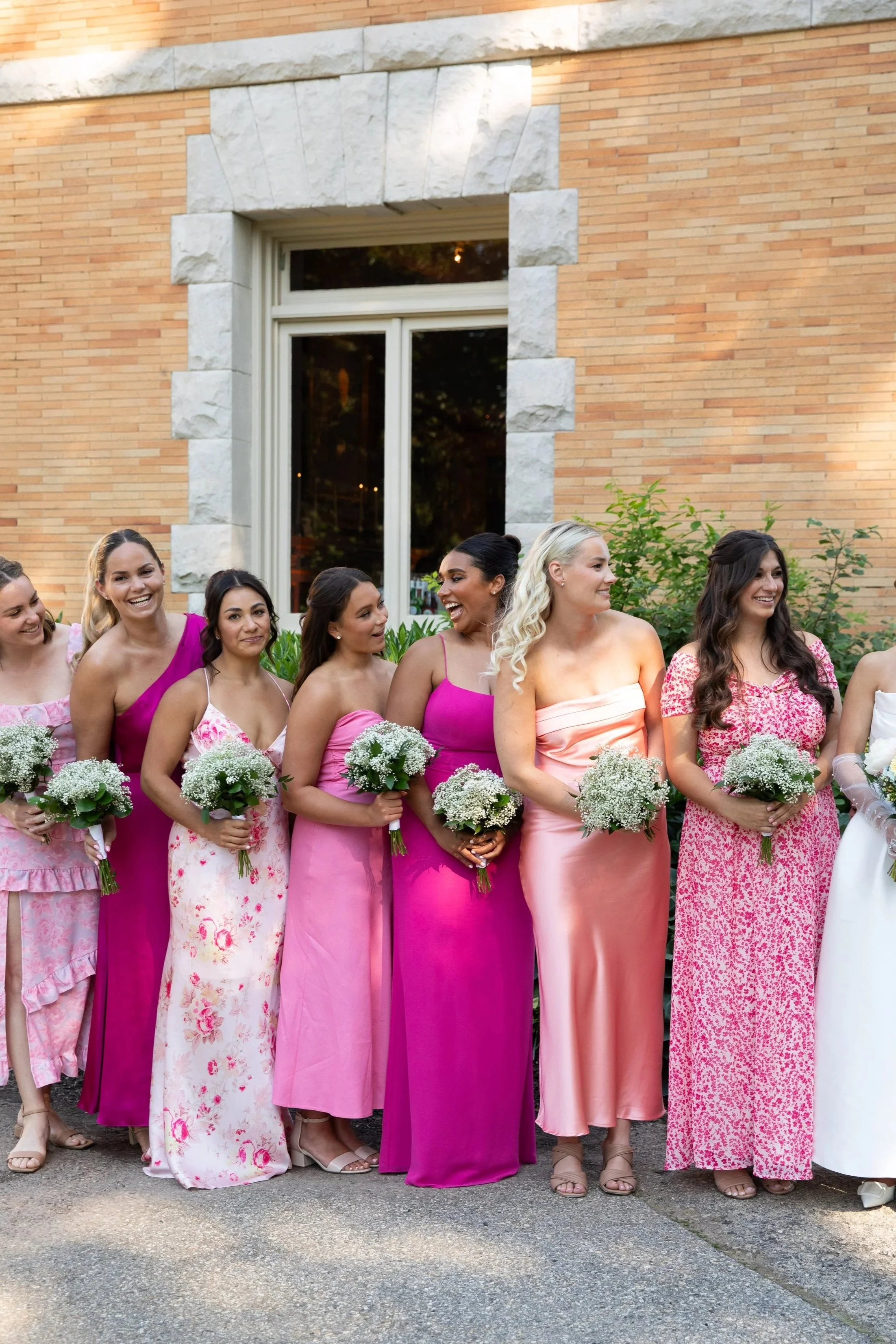 A group of women dressed in pink and floral dresses holding bouquets, standing outside in front of a brick building with a large window, during a celebration or event, possibly a wedding.