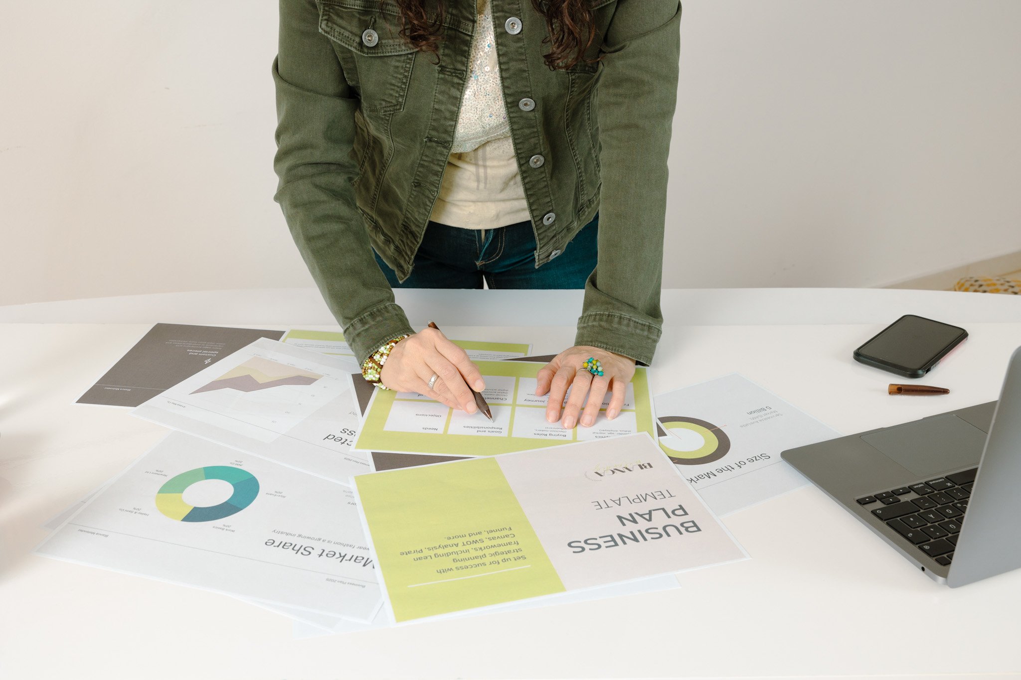 A person in a green jacket and bracelets is standing at a white table, organizing and analyzing various business and marketing reports and charts. Expert personal brand building