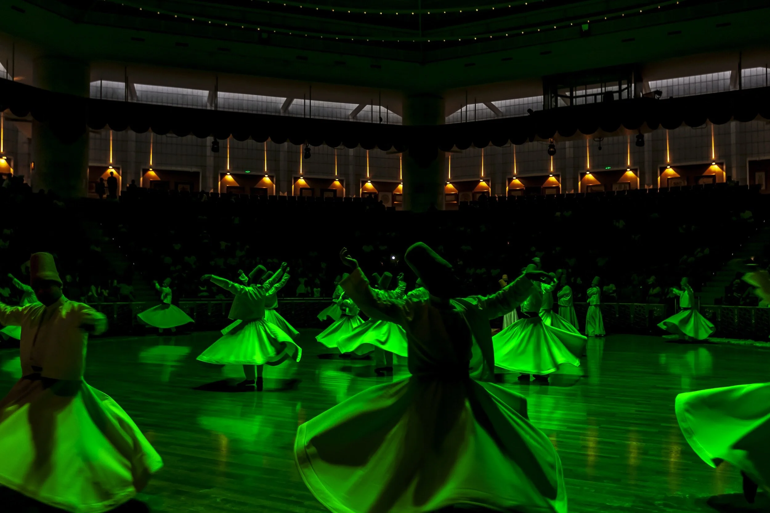 A group of dancers performing a traditional dance in a concert hall with green lighting and a wooden floor.