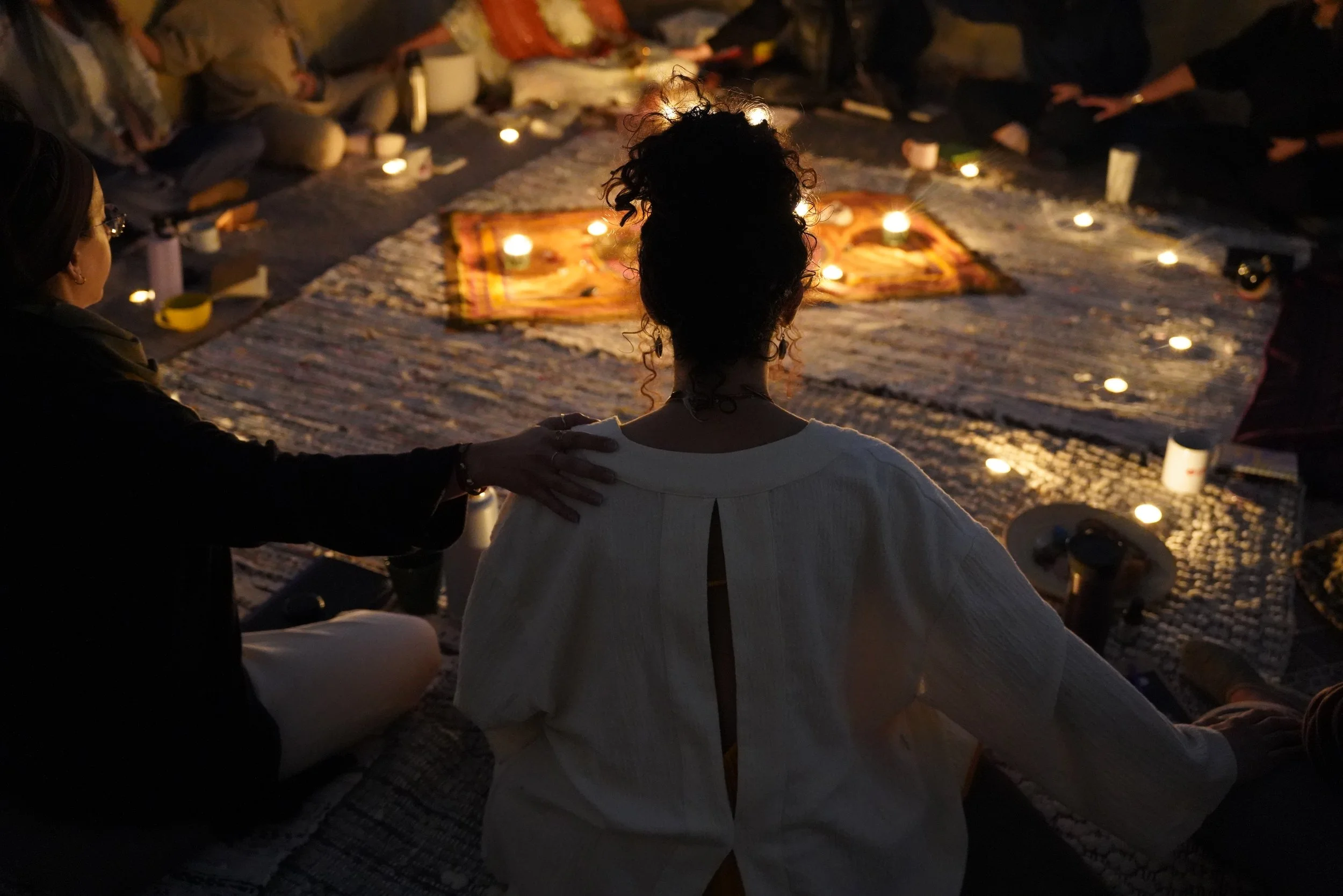 People sitting in a circle around lit candles on a blanket during a nighttime gathering or ceremony.