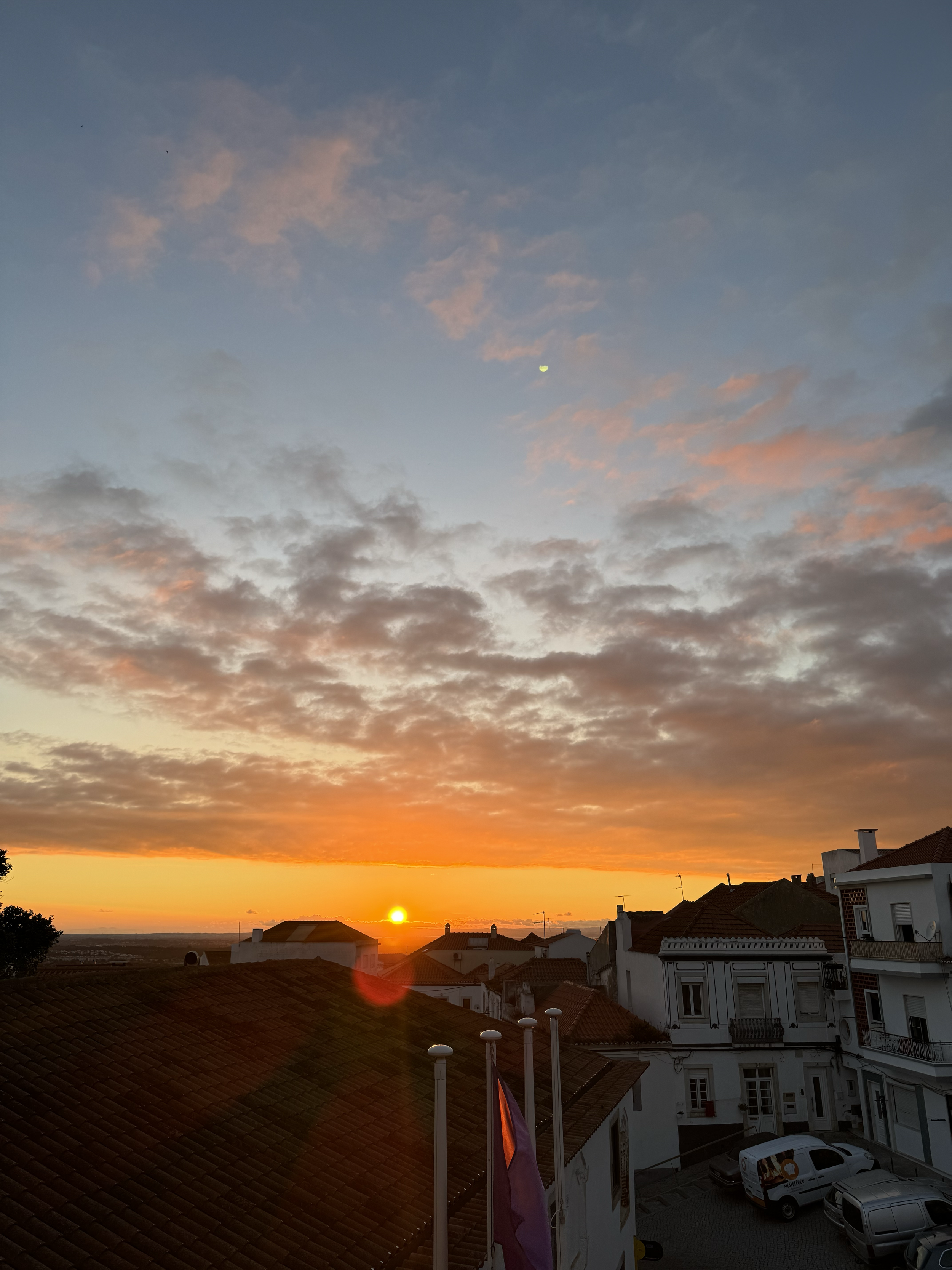 Sun setting over rooftops in a residential neighborhood, with a partly cloudy sky and a crescent moon visible.