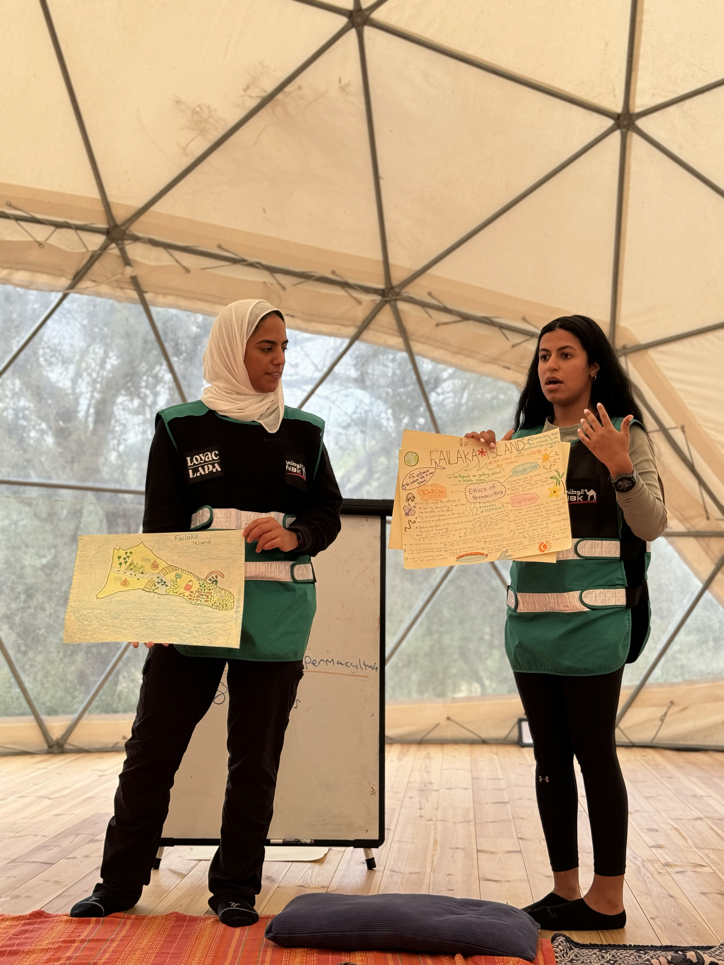Two women giving a presentation inside a geodesic dome. One woman, wearing a hijab, holds a diagram of Fakalaka Island. The other woman speaks and holds a large speech bubble poster with information and drawings about Fakalaka Island.