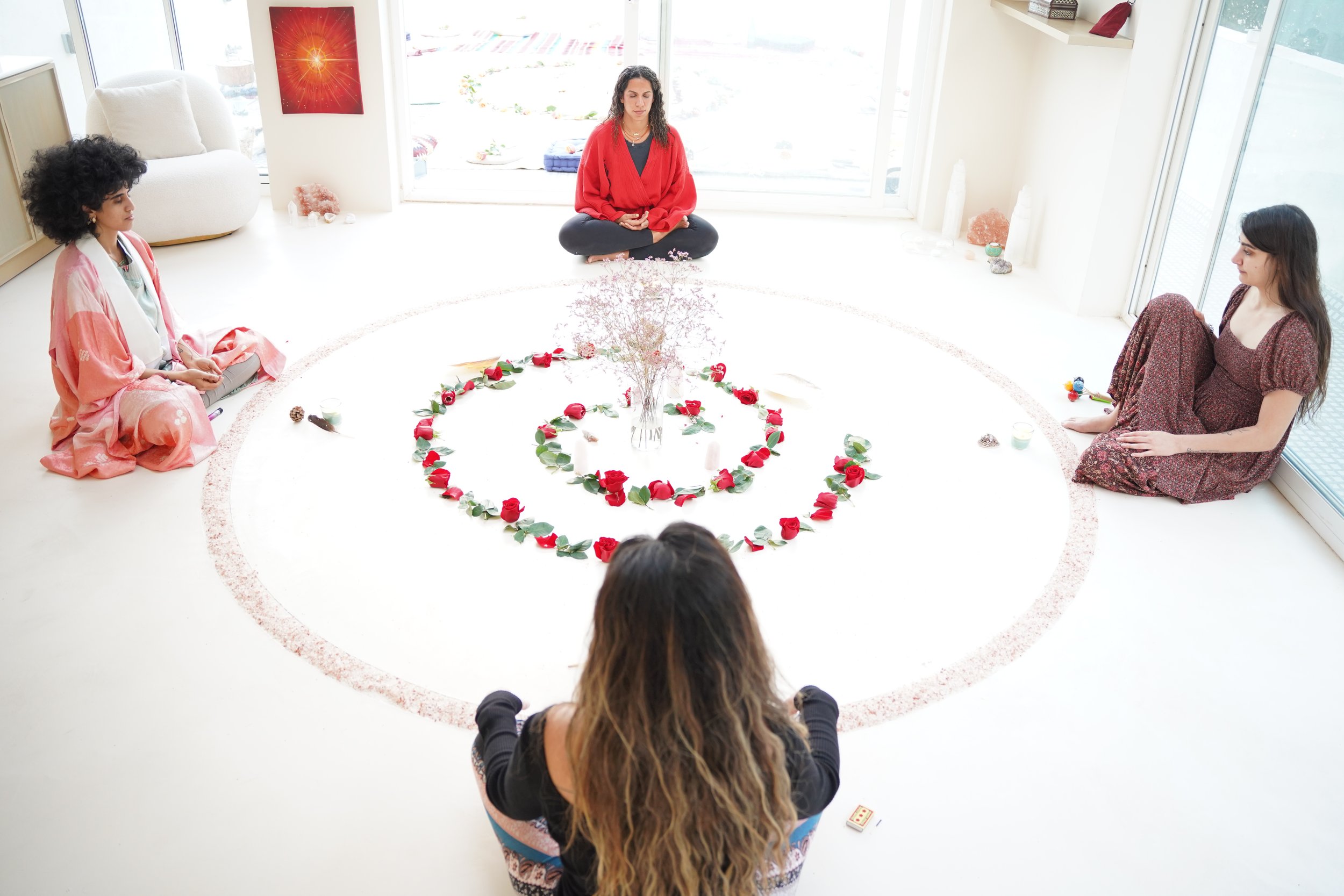 Four women meditating in a circle around a floral arrangement on a white floor in a bright room with large windows.