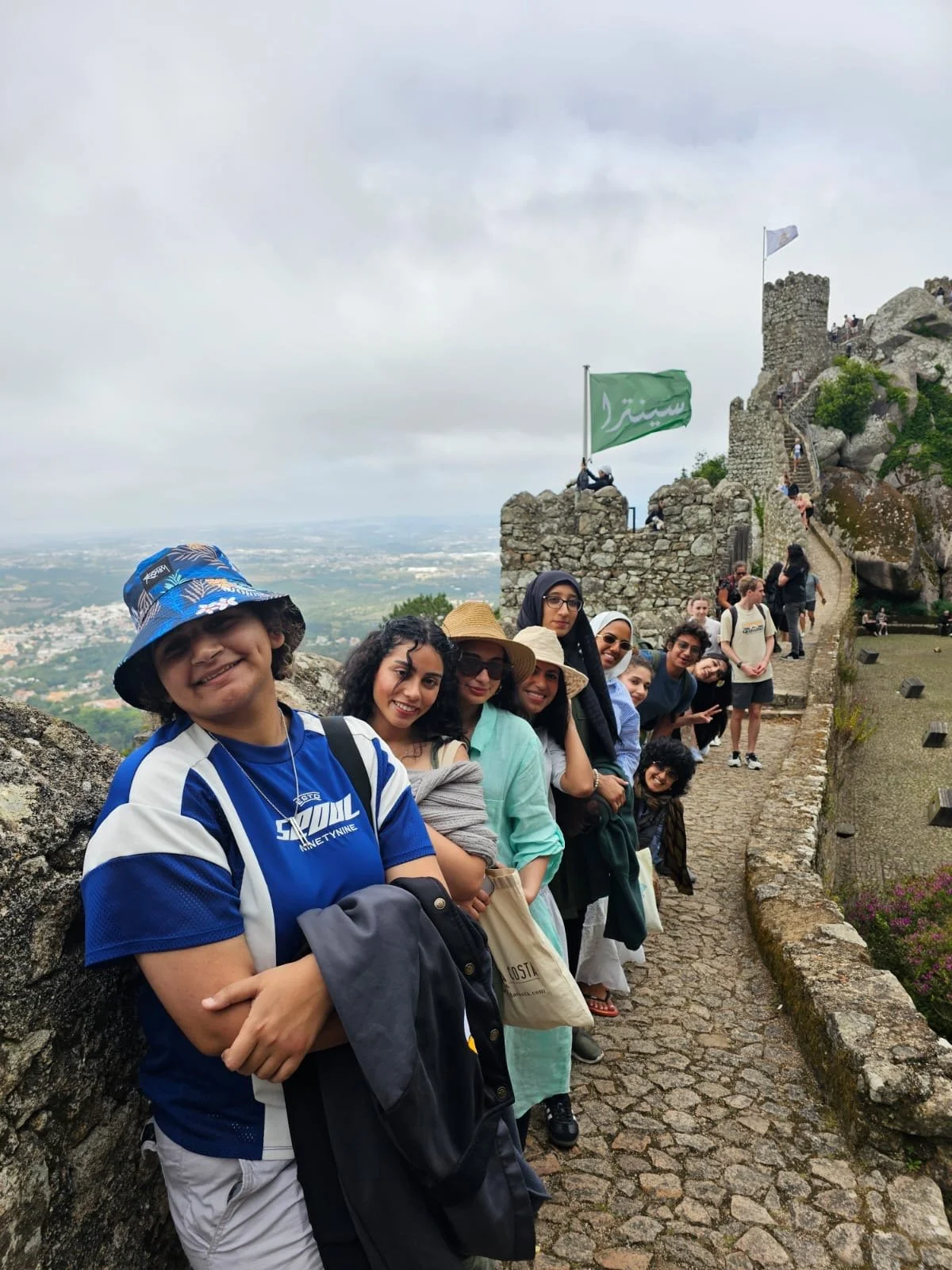 Group of people standing in a line on a stone pathway at a castle, with a cloudy sky and distant landscape in the background.