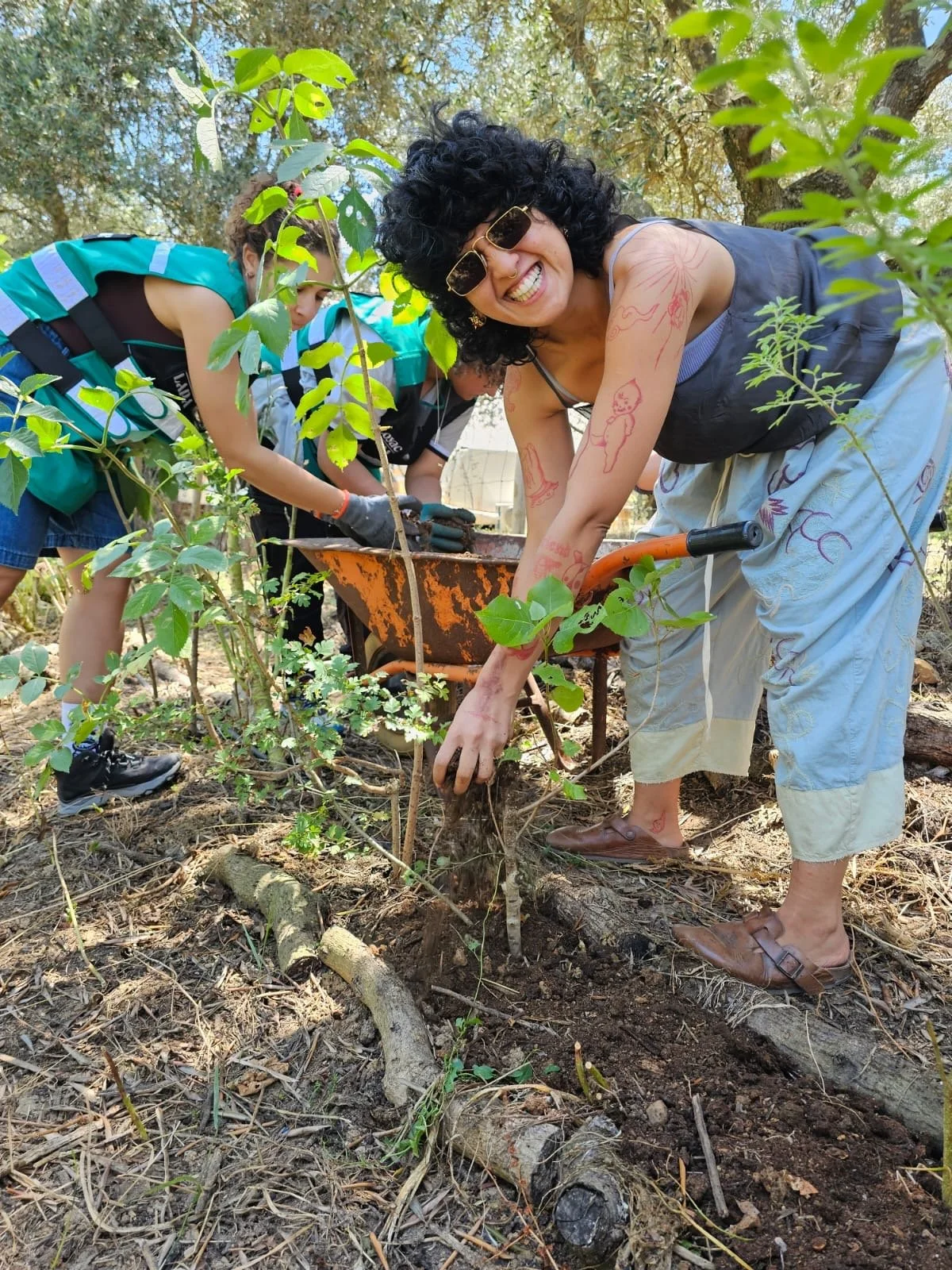 A woman planting a small tree in soil, smiling at the camera, surrounded by children helping with gardening outdoors.