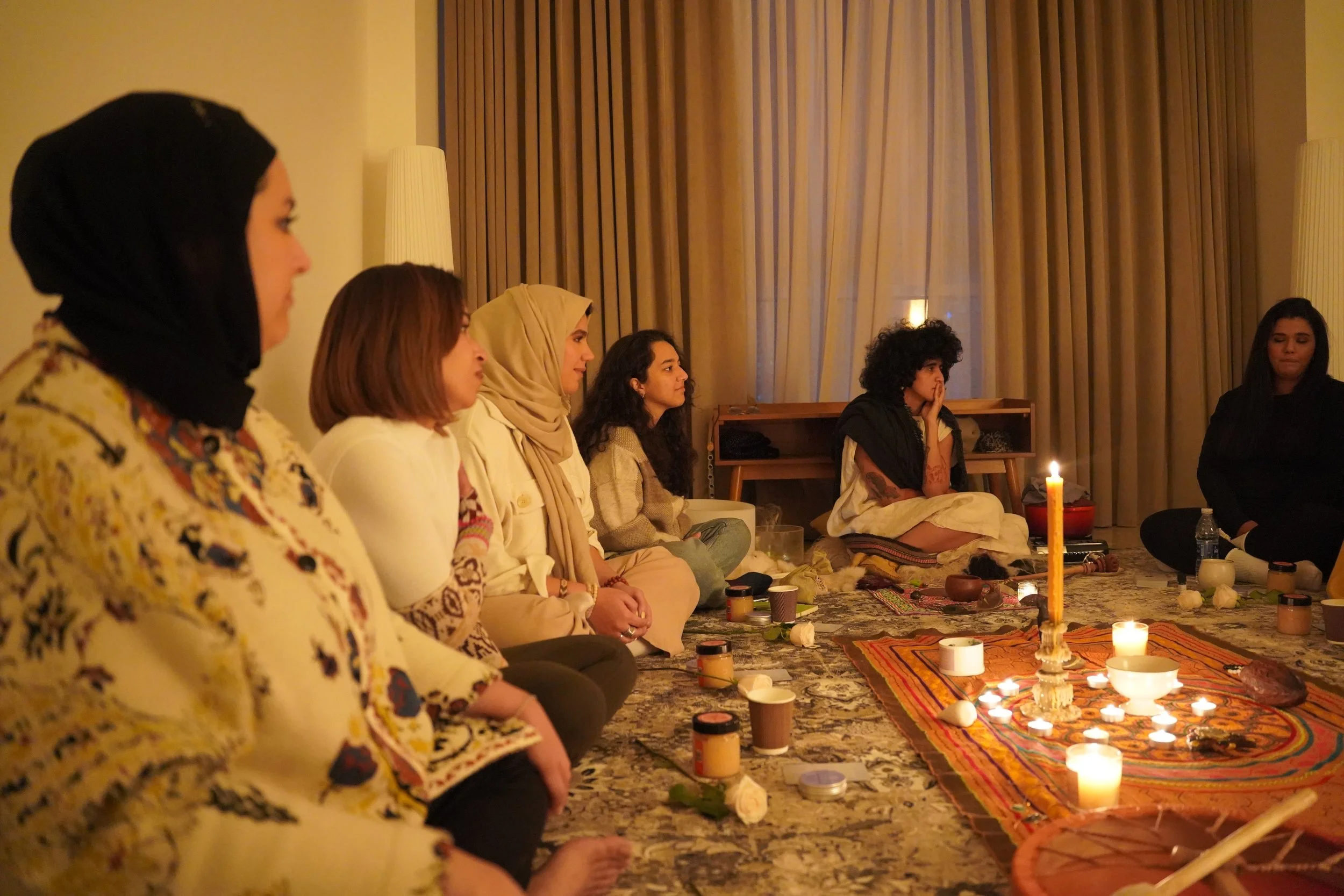 A group of women sitting cross-legged on the floor during a candlelit spiritual or meditation gathering in a softly lit room with beige curtains.