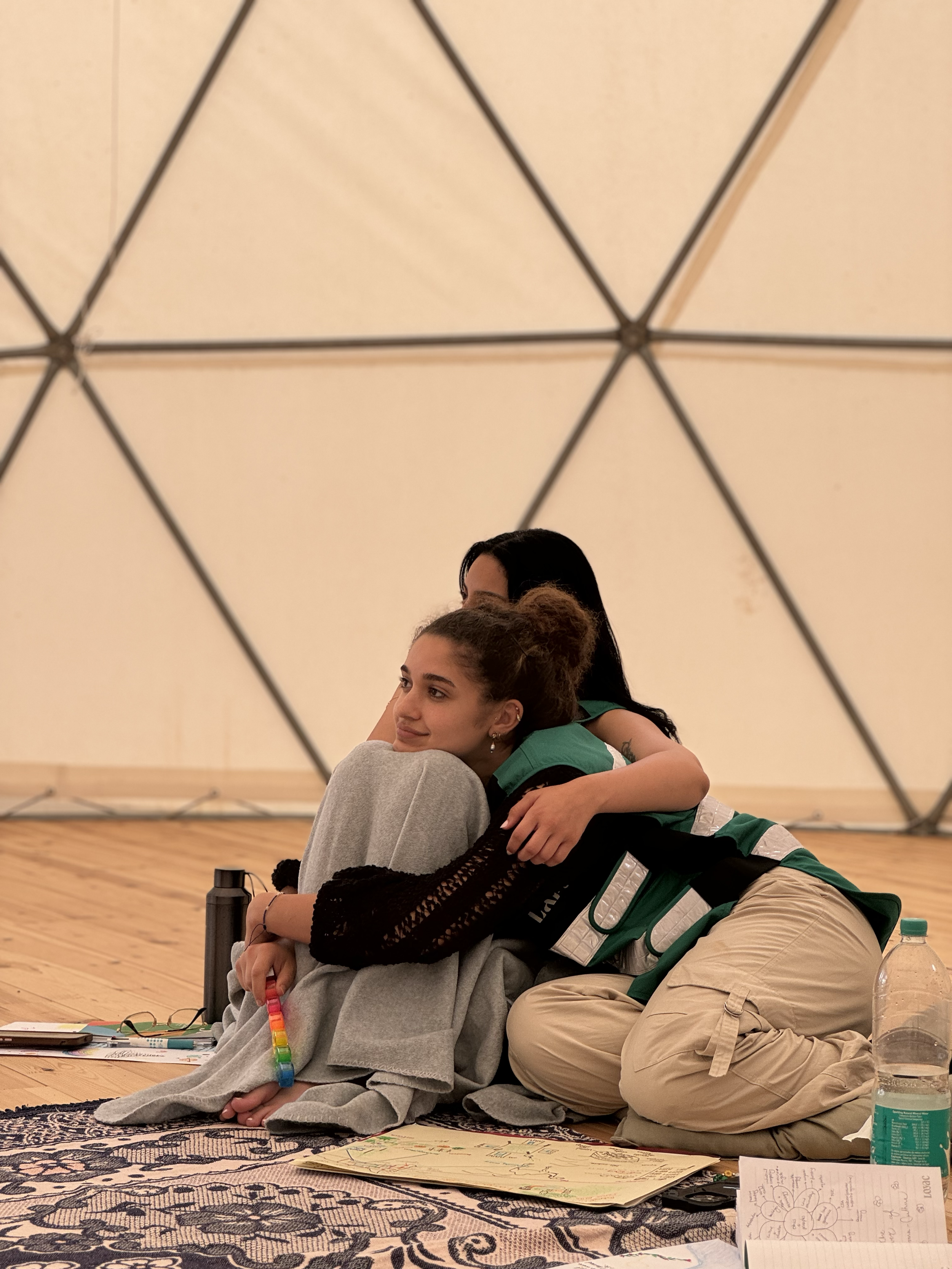 Two young women hugging. They are sitting on a patterned rug at a workspace with papers, a water bottle, and writing utensils around them.
