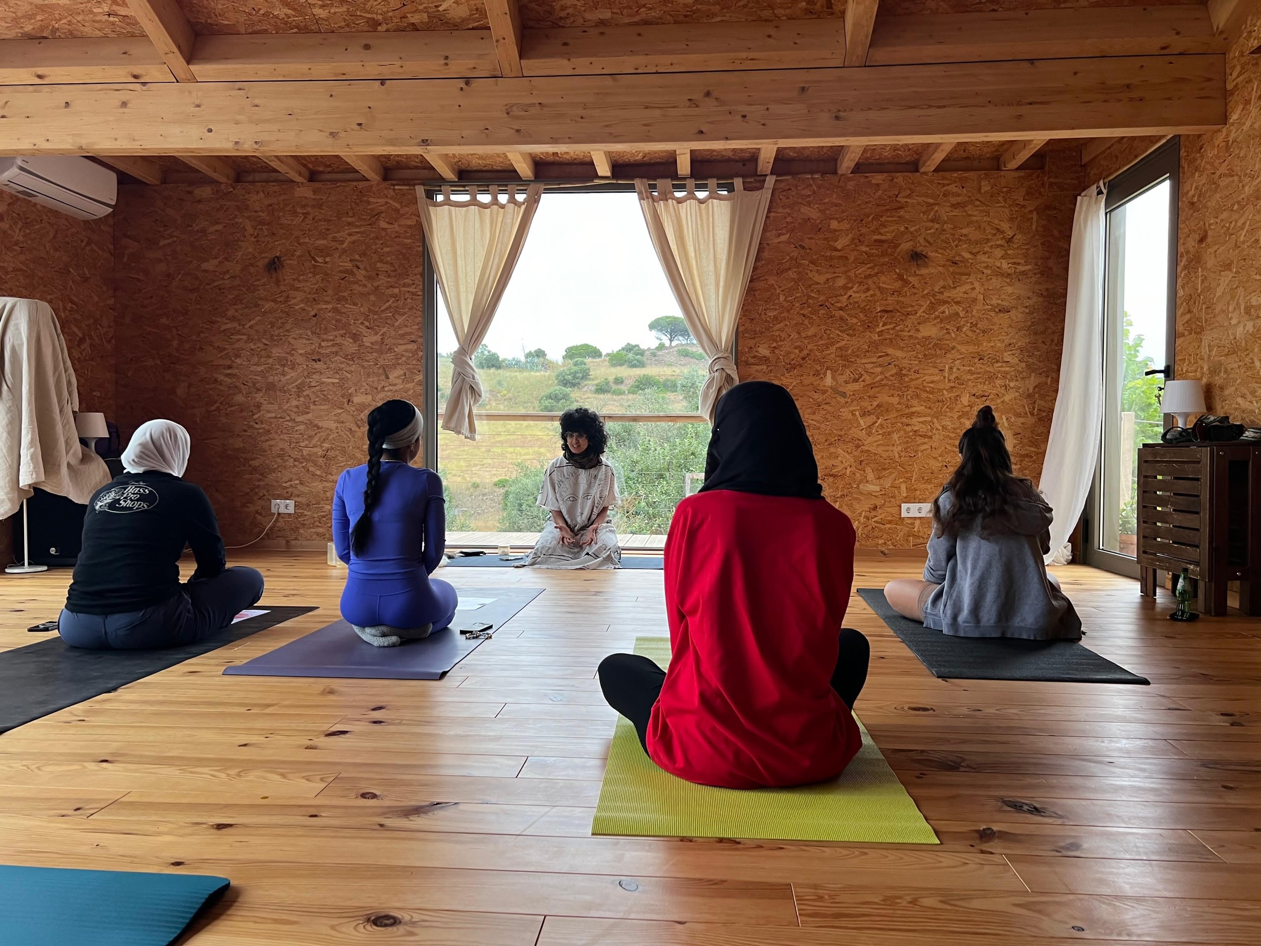 A group of people participating in a yoga or meditation class inside a wooden room with large windows, a scenic outdoor view, and a instructor sitting on the floor facing the class.