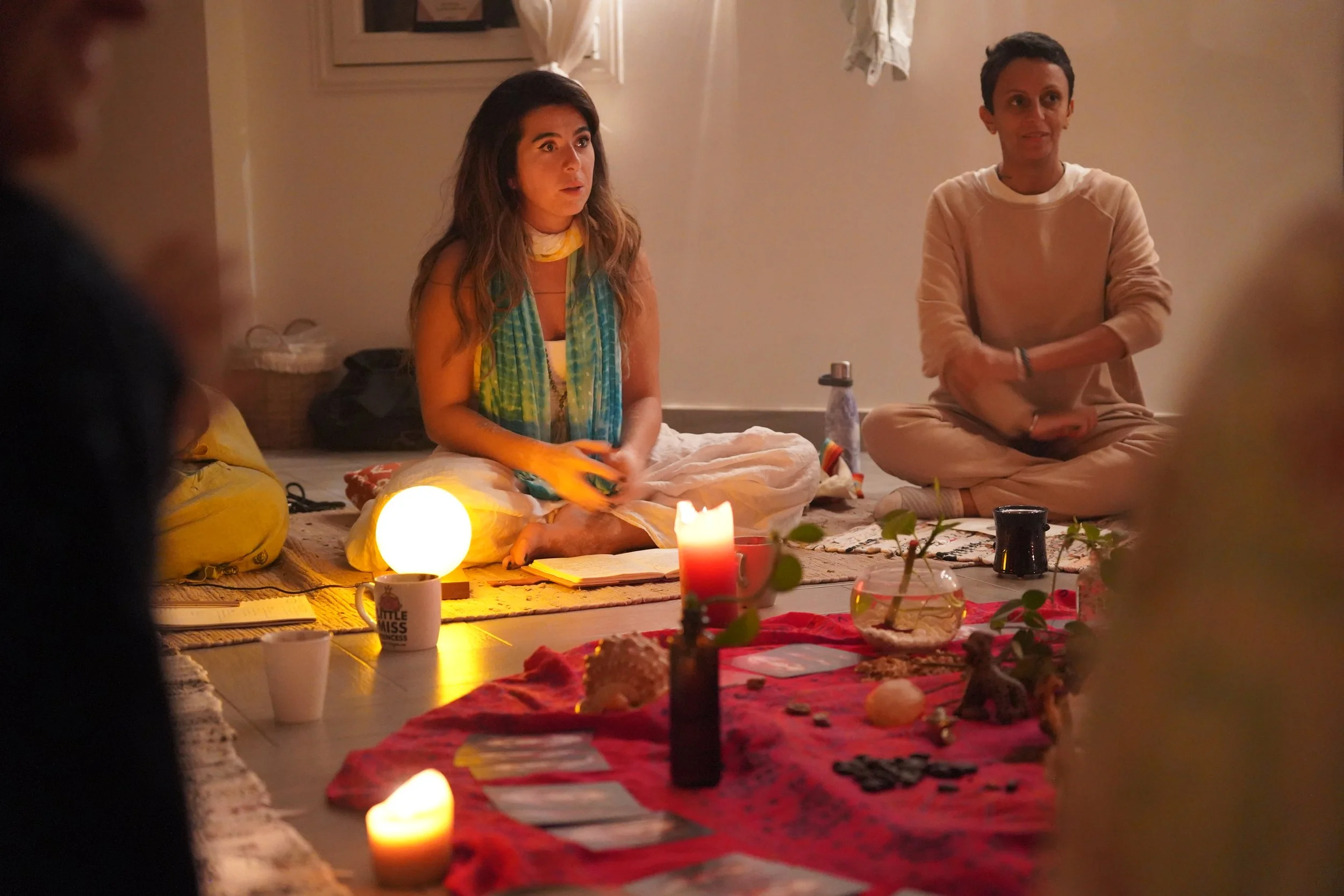 People sitting on the floor participating in a spiritual or meditation session in a dimly lit room with candles, plants, and spiritual objects.