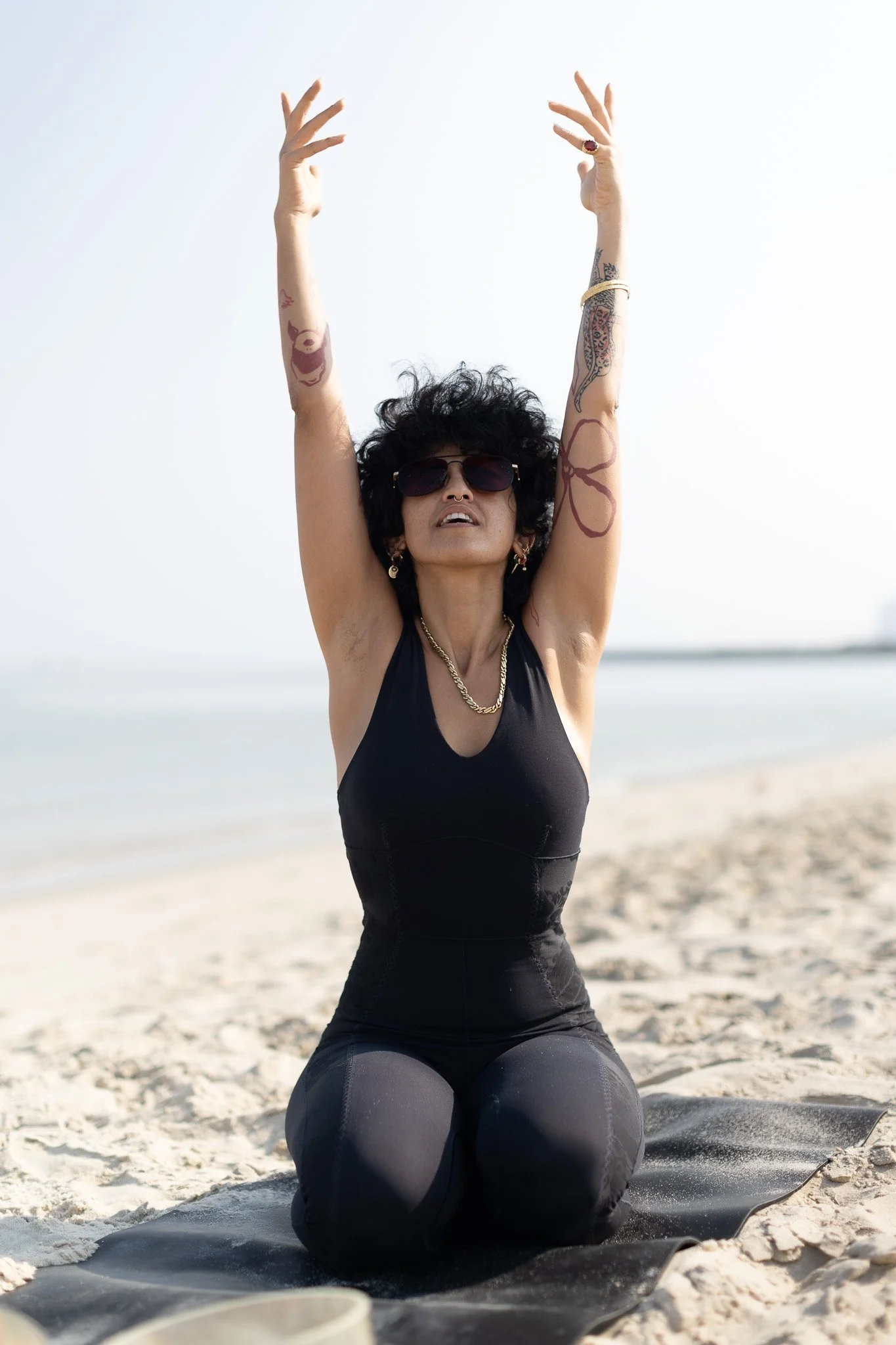 Woman with tattoos and sunglasses practicing yoga on the beach, kneeling on a mat with arms raised overhead.