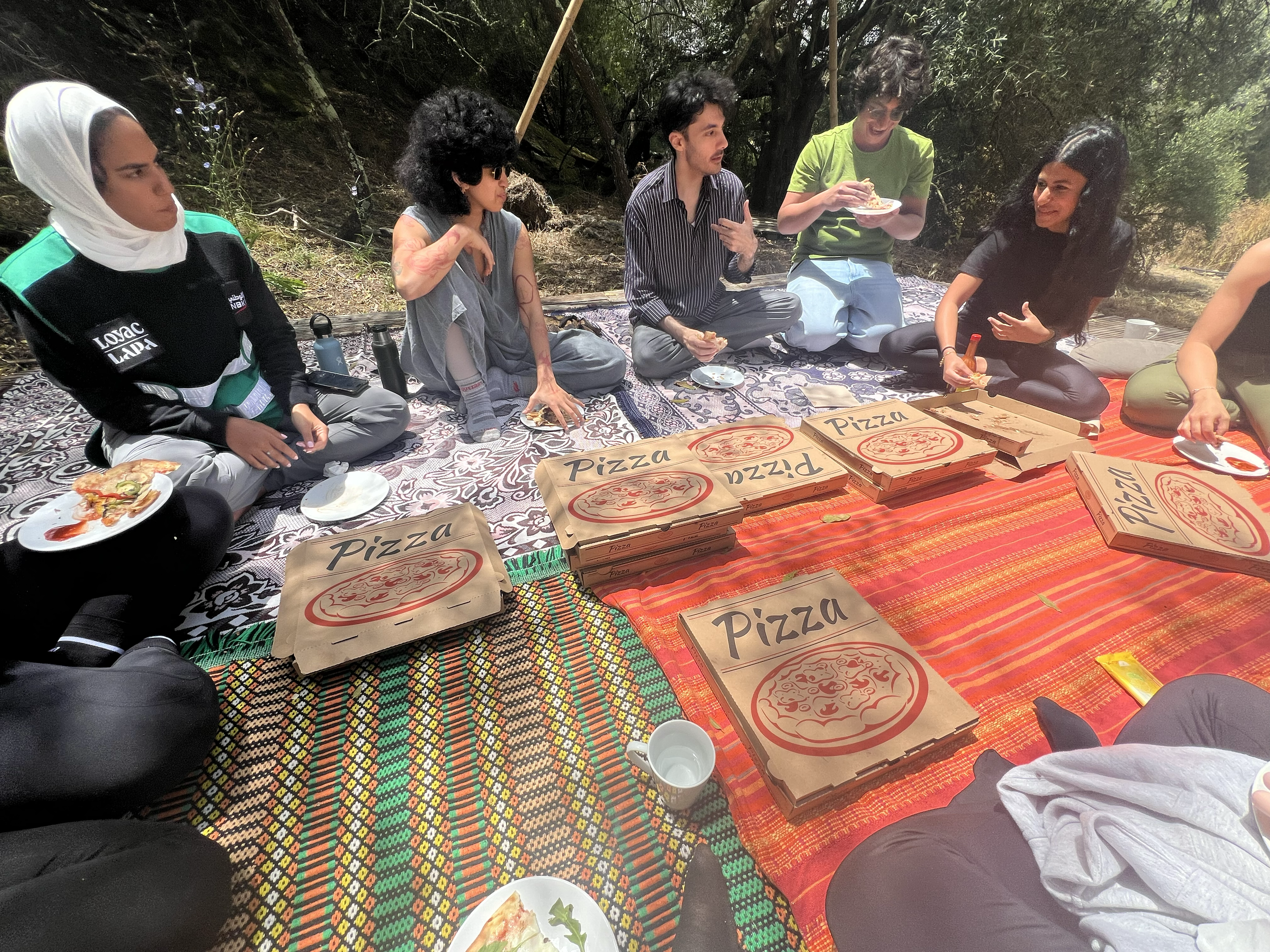 A group of people sitting on colorful blankets outdoors in a circle, with several pizza boxes in the center, some eating and some talking.