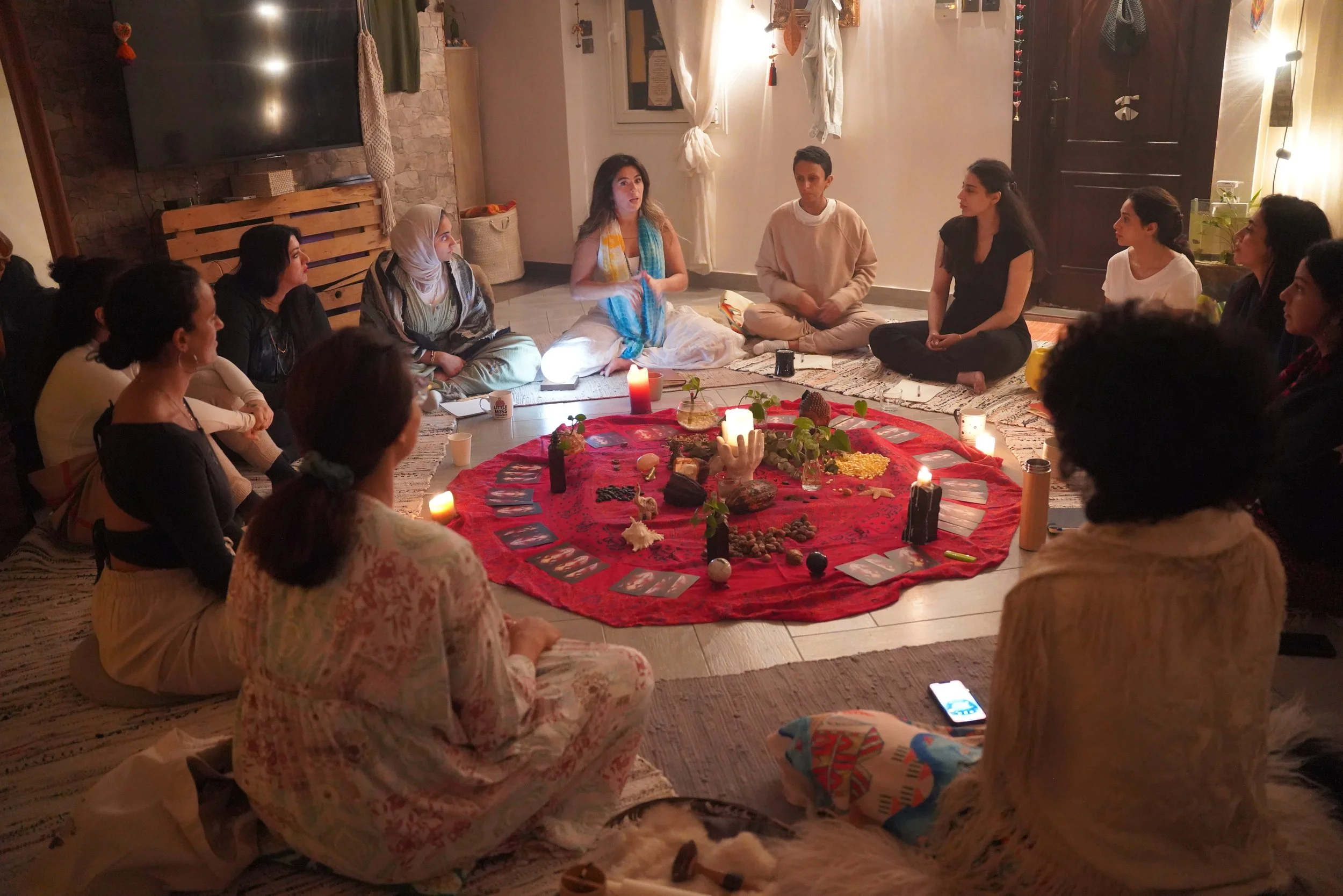 A group of people sitting on the floor in a circle around a red cloth with candles, crystals, and plants inside a cozy, dimly lit room, probably engaged in a spiritual or meditation gathering.