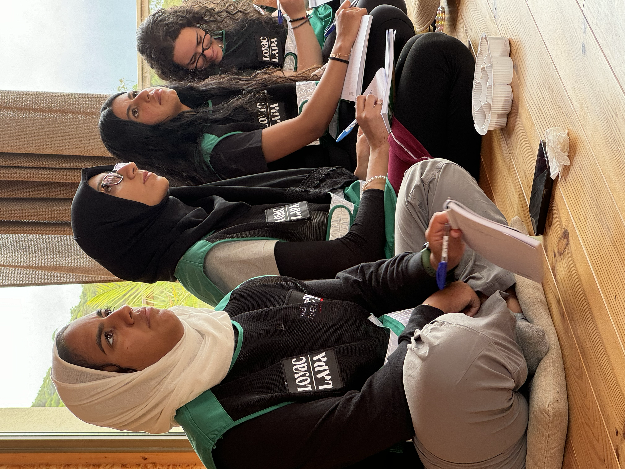 Four women sitting on the floor, holding notebooks and pens, engaged in a discussion or meeting. The woman in the front is wearing a beige hijab and glasses, while others are wearing black and green shirts. They are seated on cushions on a wooden flo