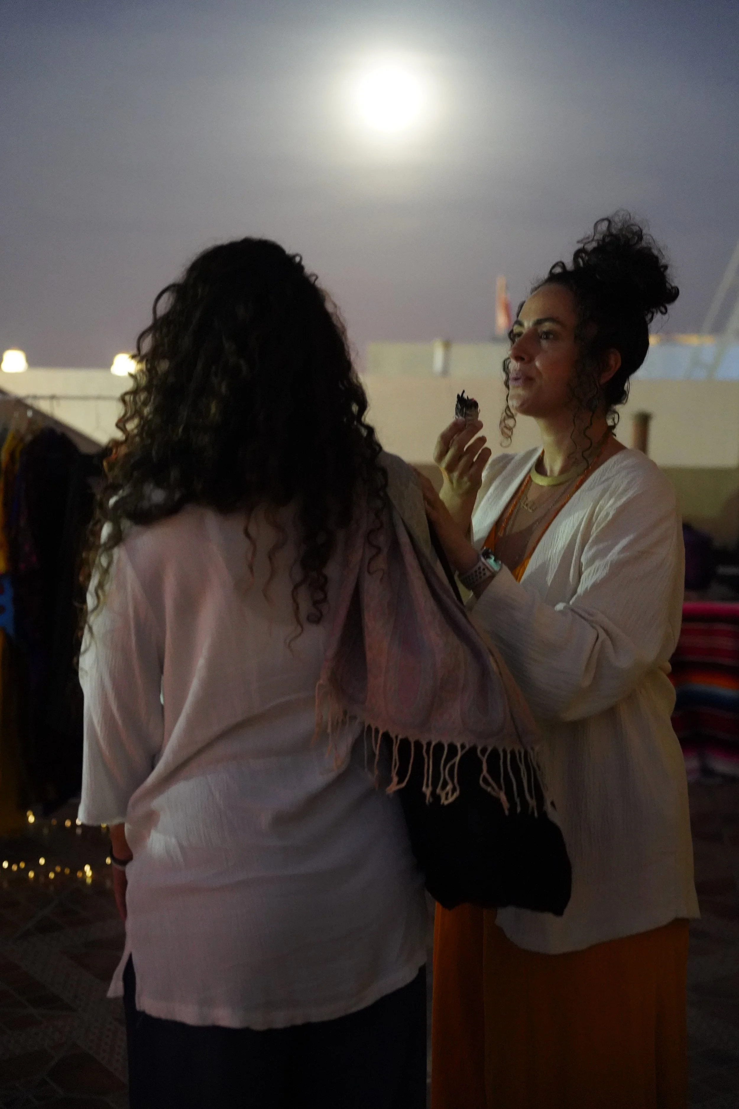 Two women are standing outdoors at night under the bright full moon. One woman is facing away from the camera, wearing a white shirt and carrying a colorful scarf or shawl. The other woman, facing the camera, with curly hair tied up, is eating a snack and wearing a white jacket over a brown dress.