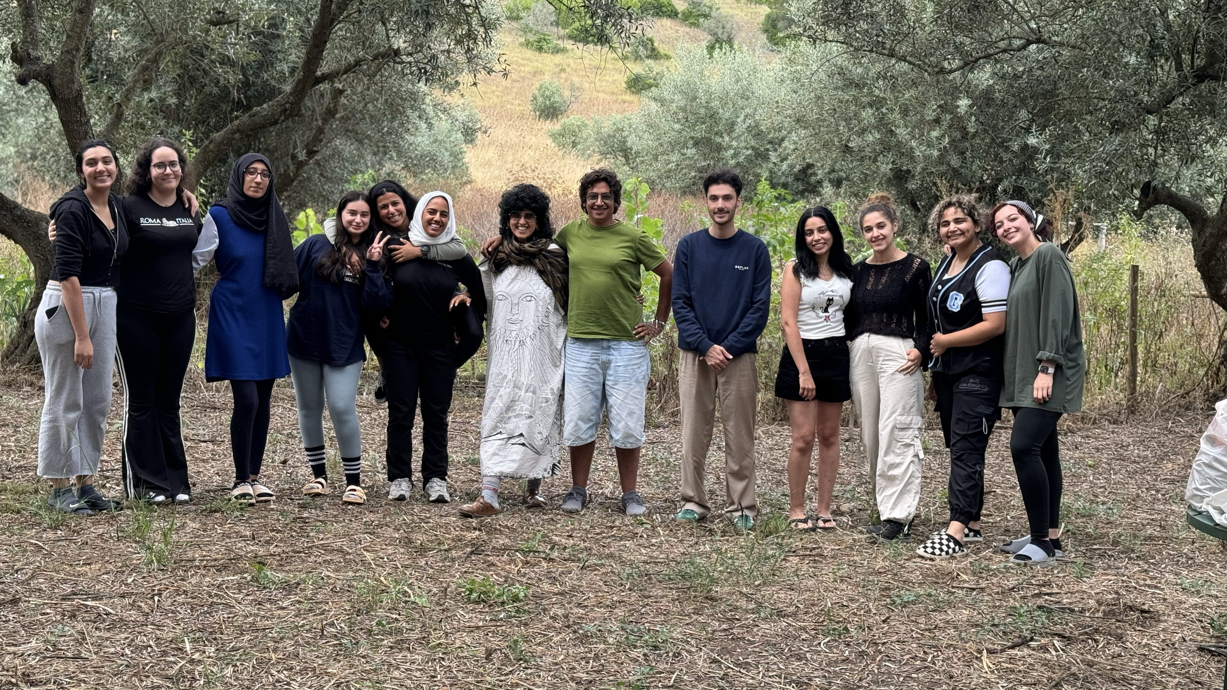 Group of fifteen people standing outdoors in a natural setting, smiling and posing together, with trees and a grassy field in the background.
