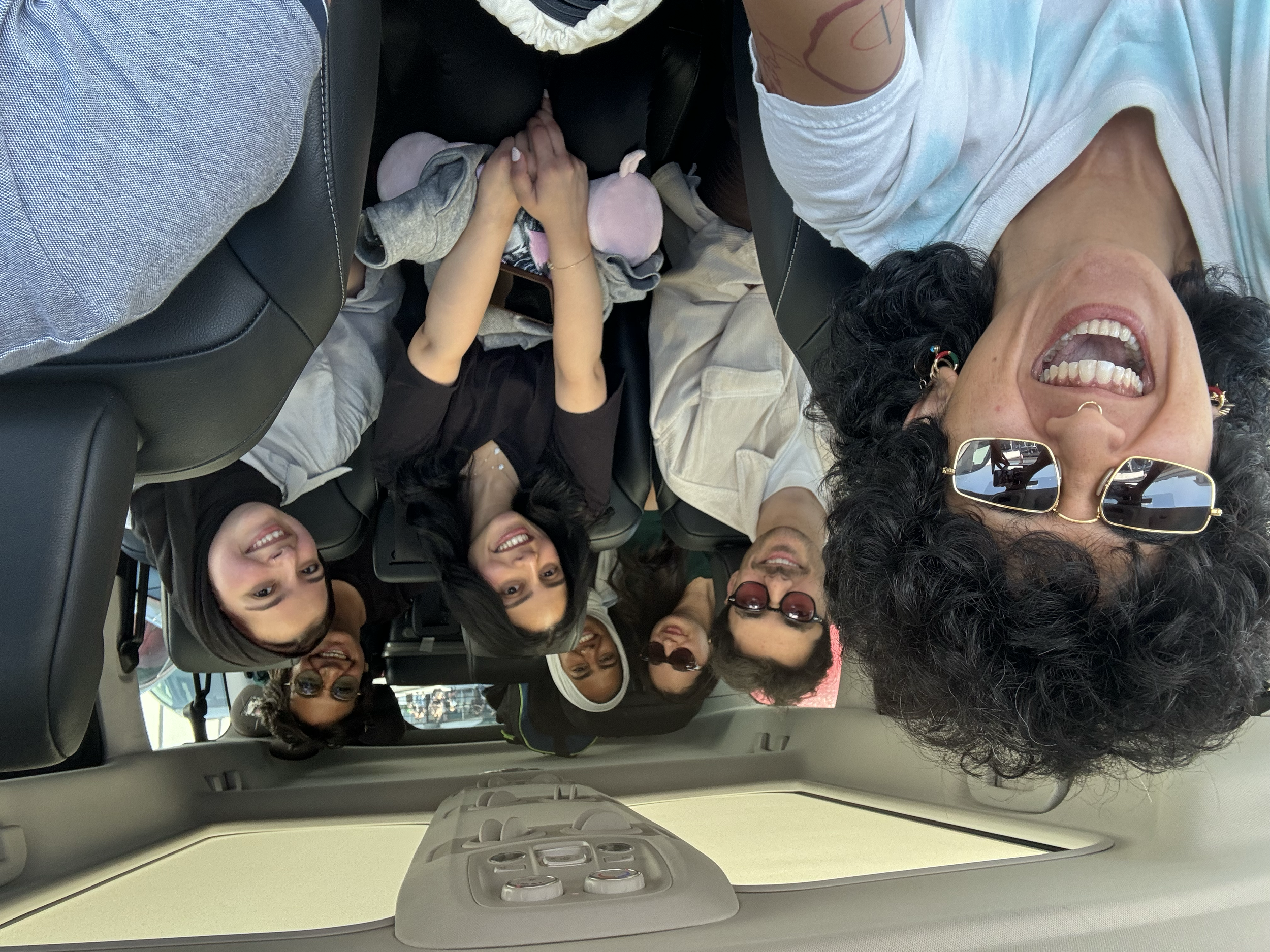 Group of women and a girl sitting in the back seat of a car, smiling and taking a selfie. One woman is wearing sunglasses and has curly hair, and the girl in the middle is holding hands with another woman.