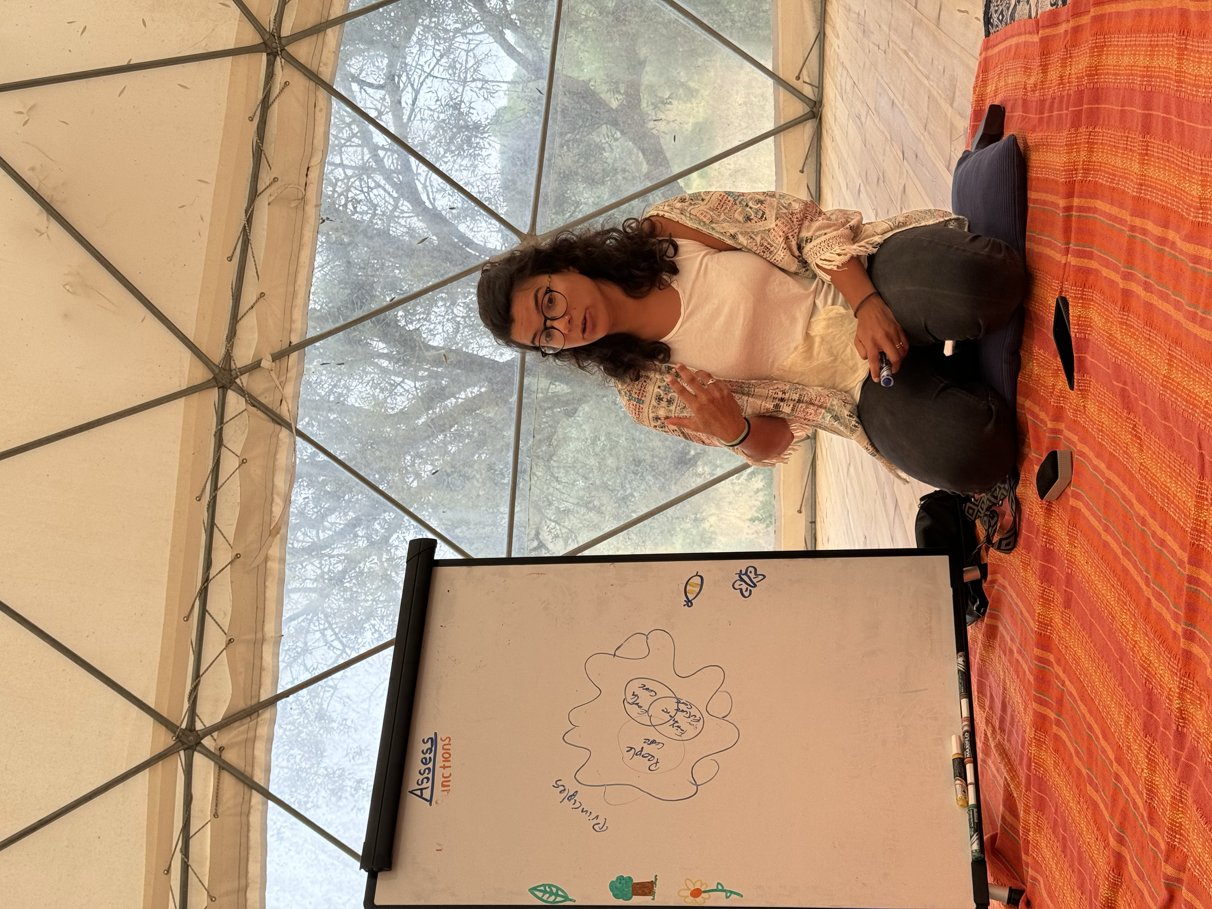 A woman sitting on a colorful tablecloth inside a tent with a whiteboard in front of her, holding a pen, making a peace sign with her right hand, and gesturing with her left. There are markers and a phone on the table.