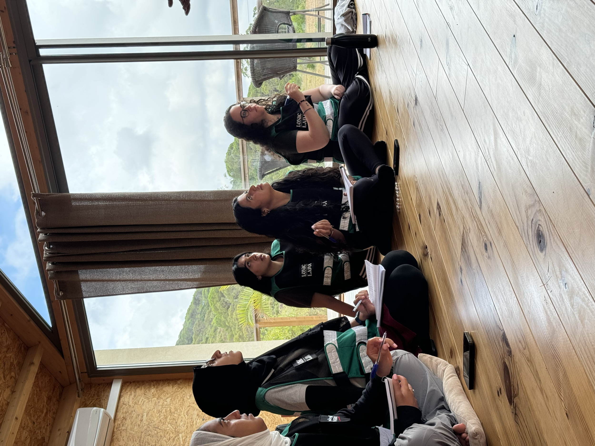 A group of four women sitting on the floor in a wooden room with large windows, some with notebooks, engaged in a discussion or meeting with a scenic outdoor view in the background.