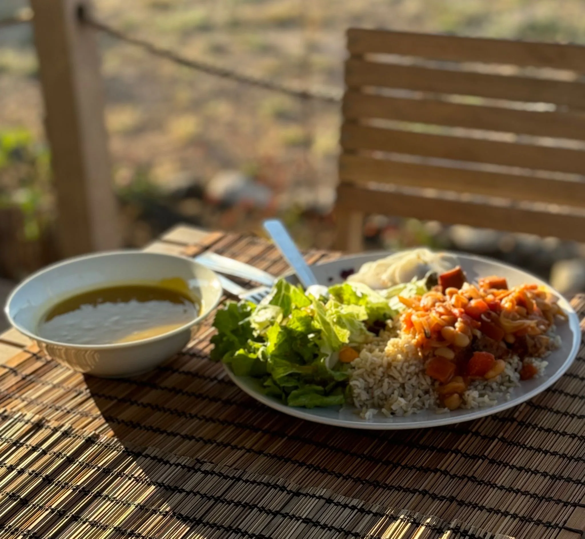 A plate of rice with vegetable stew, a side of lettuce, a scoop of mashed potatoes, and a bowl of soup on a bamboo mat outside.