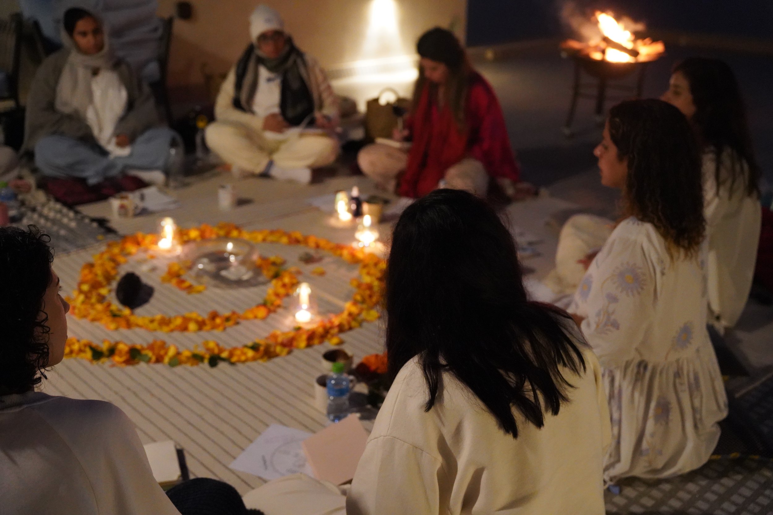 People sitting on the ground around a sacred fire with flower garlands, candles, and ritual items during an evening ceremony.