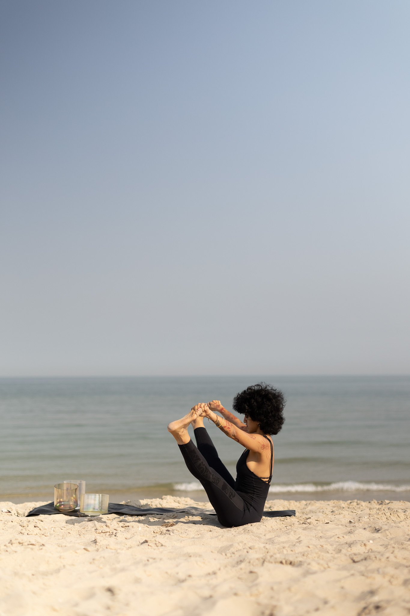 A woman practicing yoga on a sandy beach by the ocean, sitting on a yoga mat with crystal singing bowls nearby.
