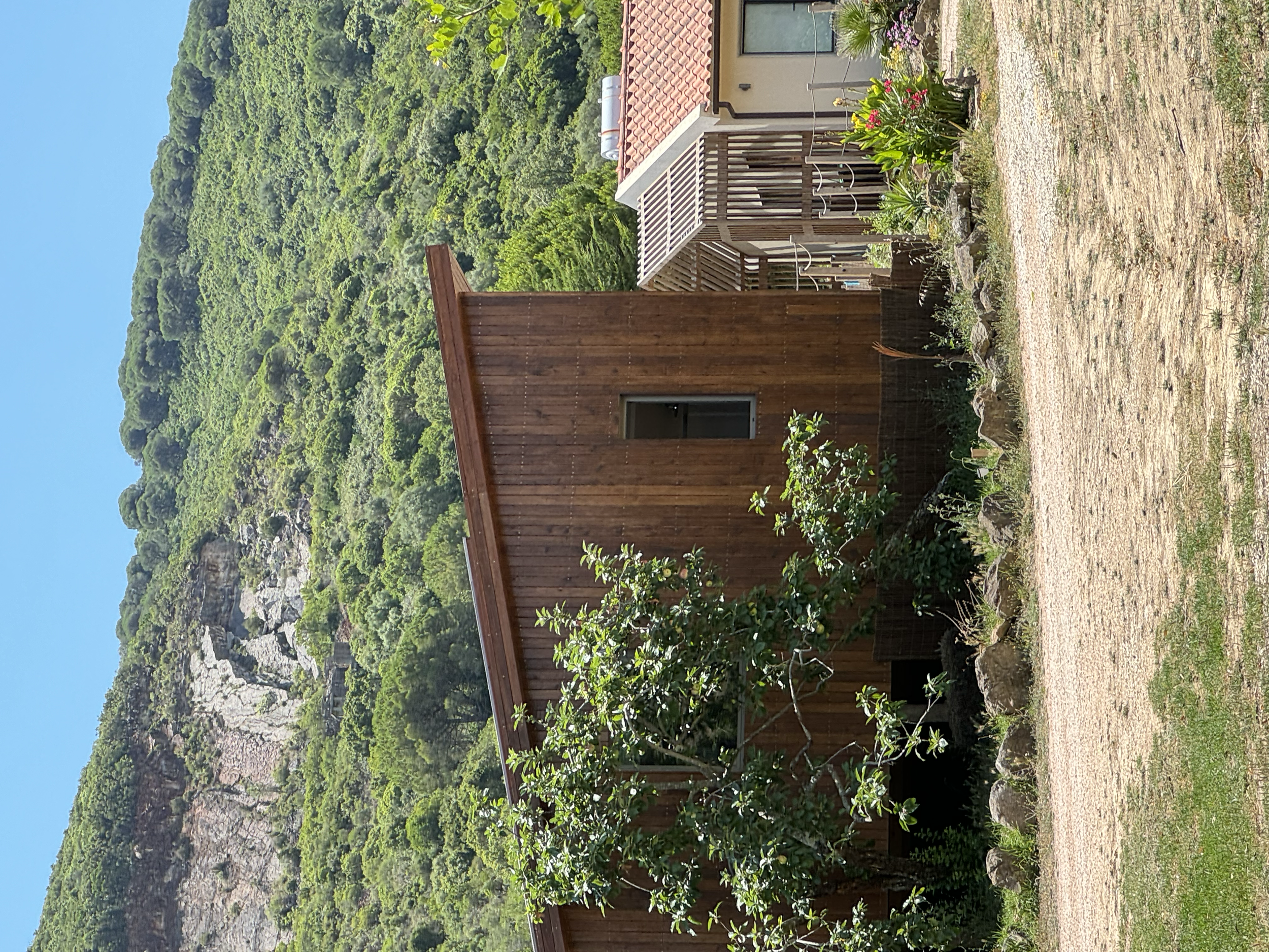 Small wooden building with a single window, surrounded by green plants and a gravel path, with a larger house and lush green hills in the background.