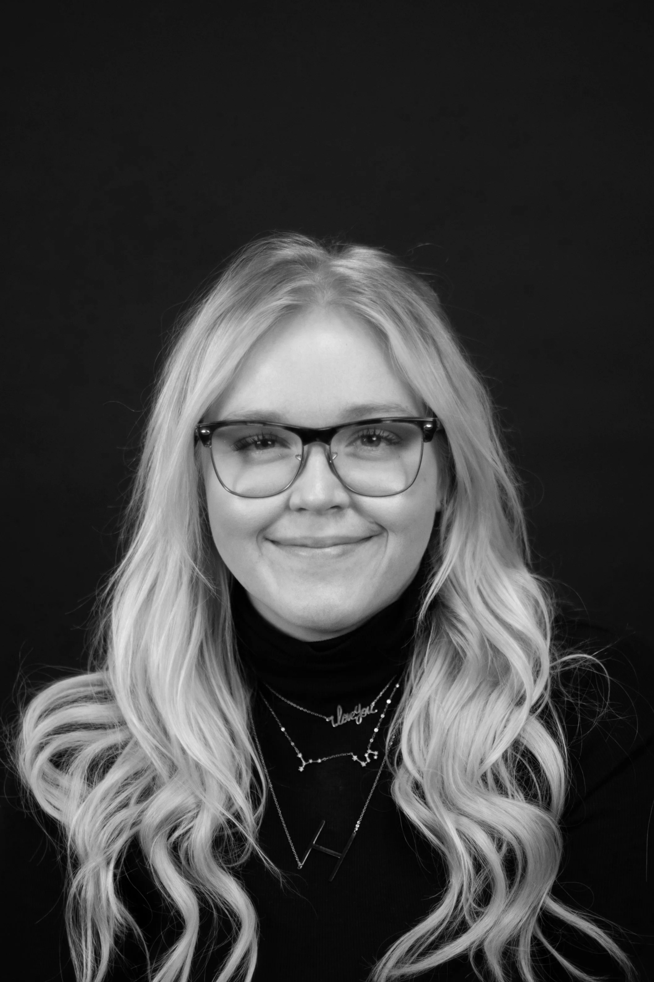 Black and white photo of a smiling woman with long, wavy blonde hair, wearing glasses, necklaces, and a turtleneck, against a dark background.