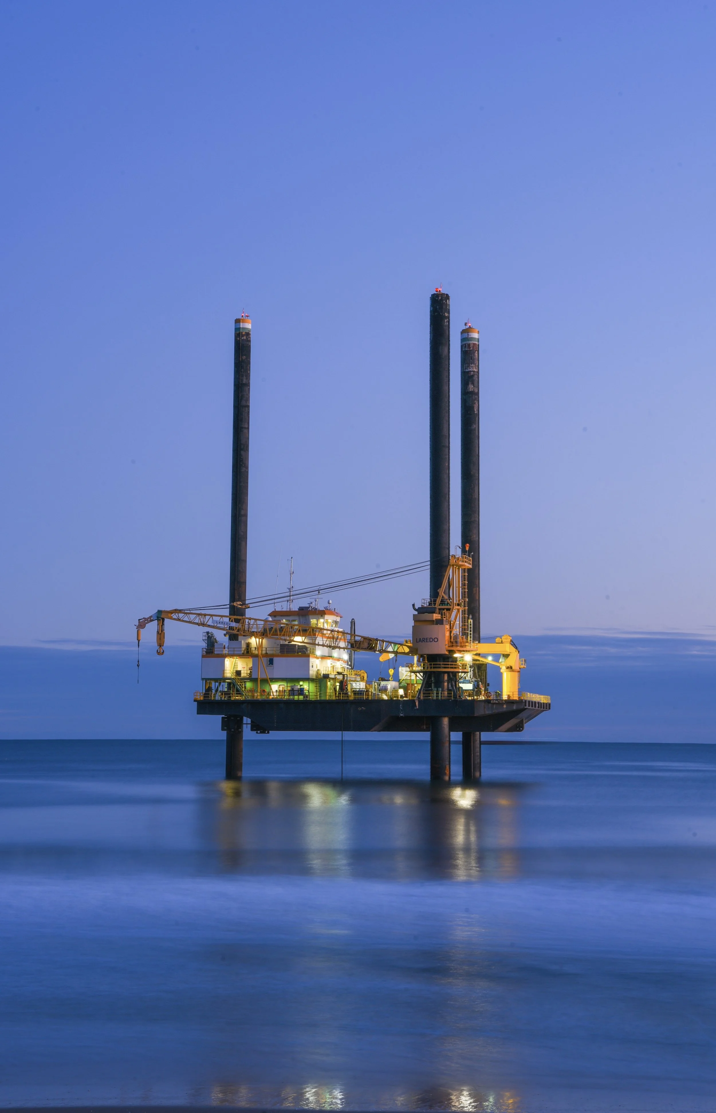 Offshore oil drilling platform illuminated at dusk with reflection on calm ocean water.