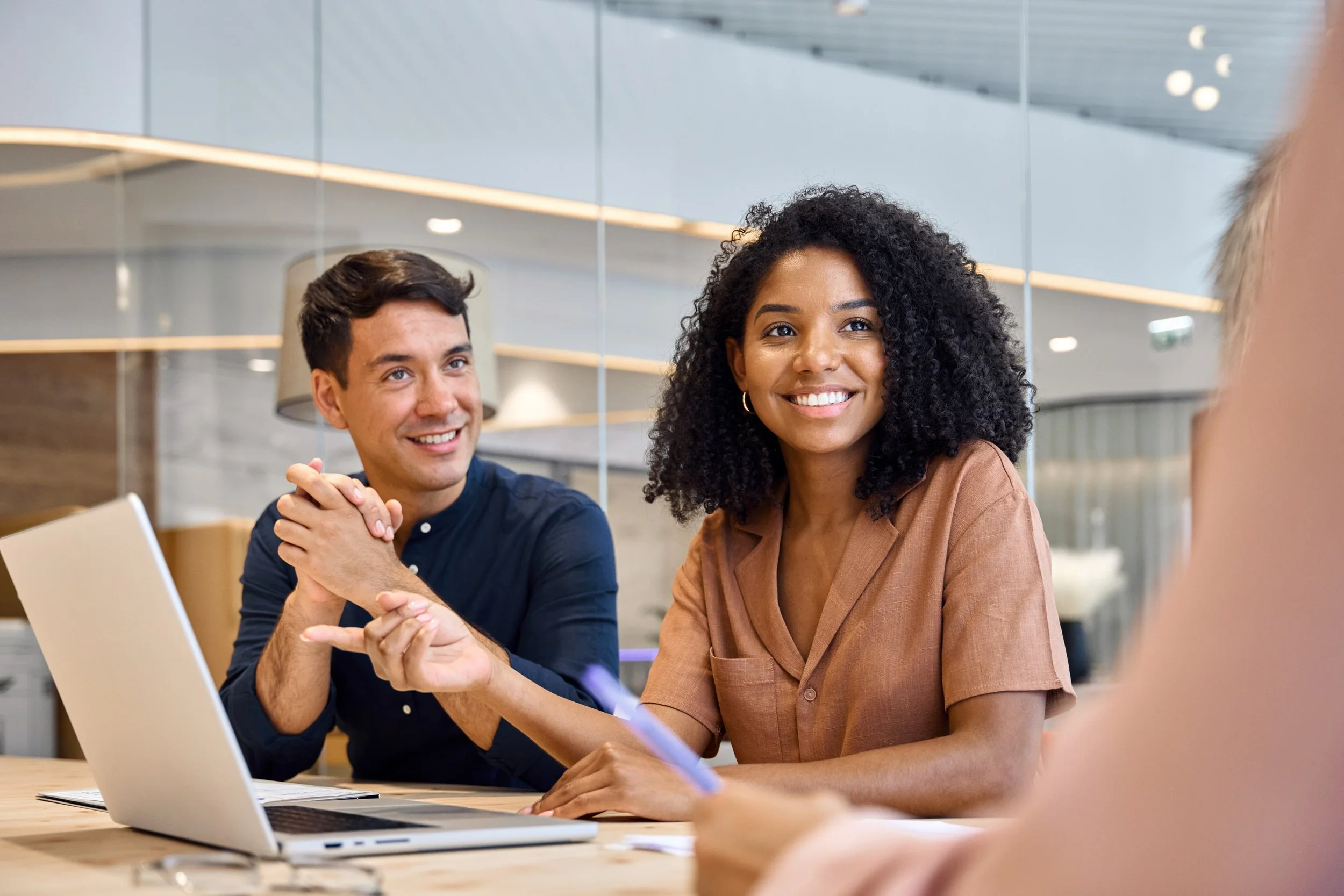 Two smiling people, a man and a woman, engaging in a conversation at a modern office. The woman, with curly black hair, is wearing a brown shirt, while the man, with short dark hair, is in a dark blue shirt. There is a laptop open on the table in front of them.