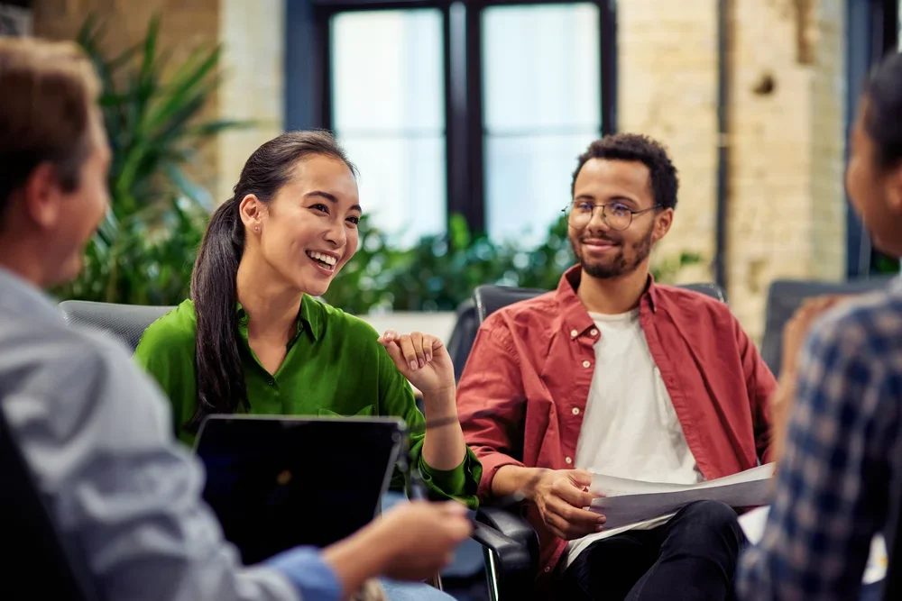 Group of four diverse young adults having a meeting or discussion in a modern office with large windows and green plants.
