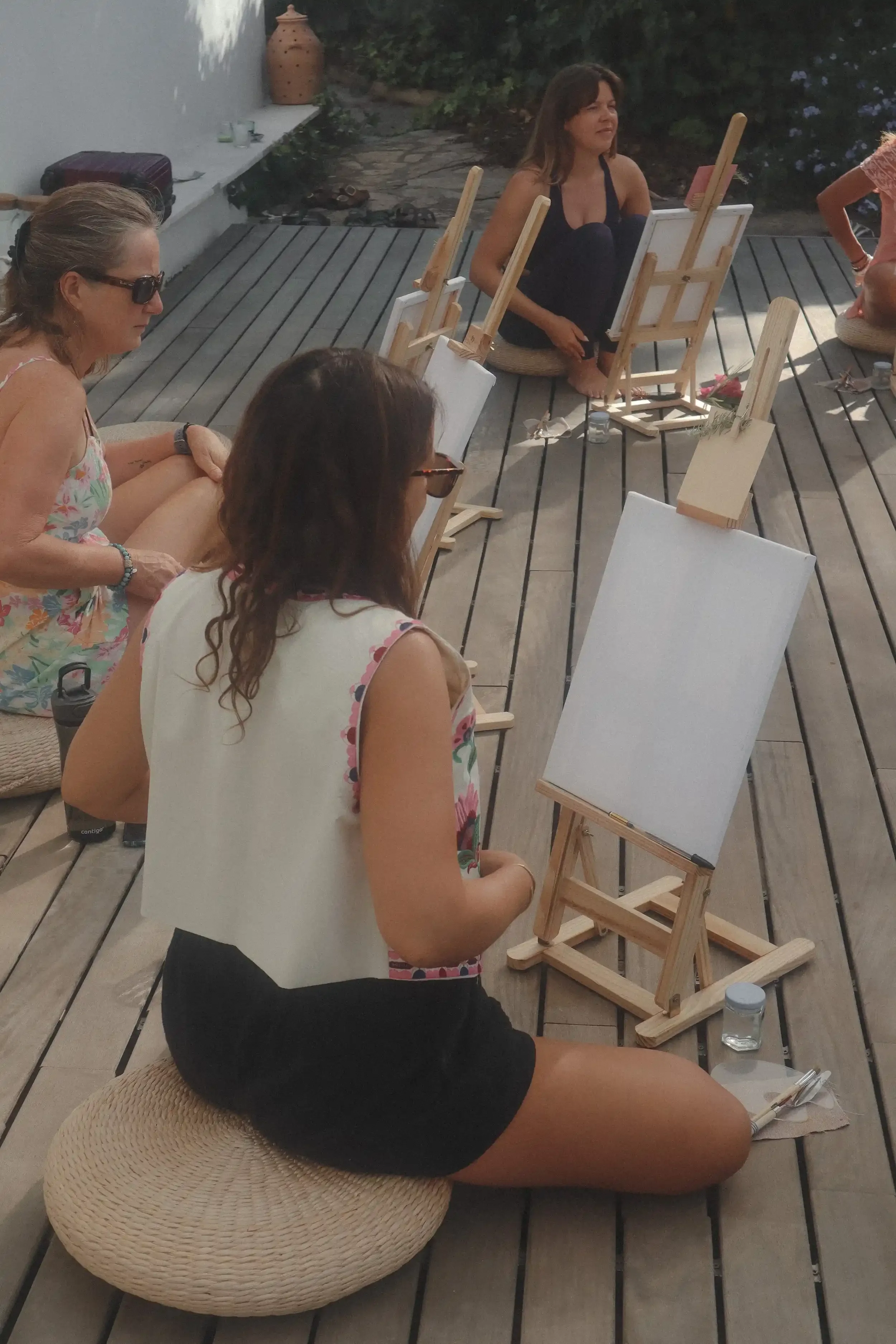 Personas participando en una clase de pintura al aire libre, sentadas en cojines, con lienzos en caballetes.