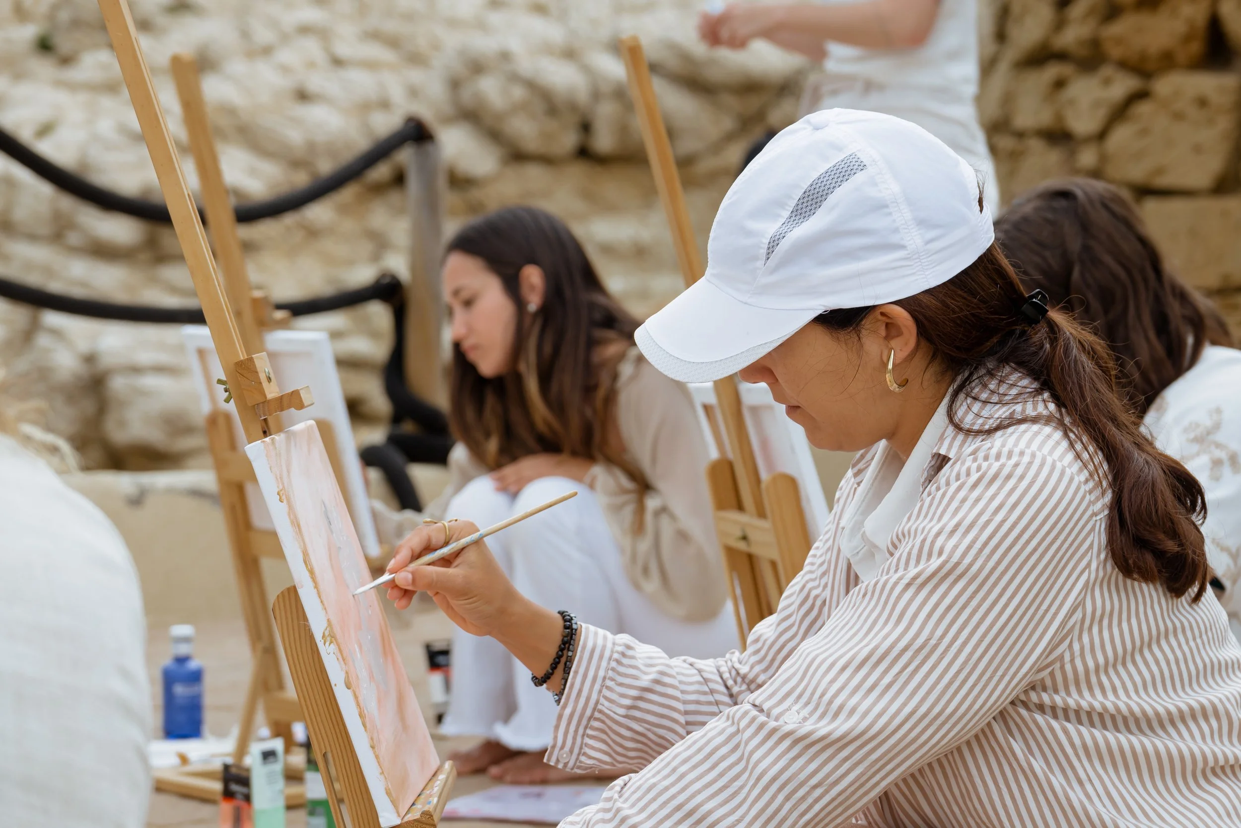 A woman wearing a white cap and striped shirt painting on a canvas outdoors with other people painting in the background.