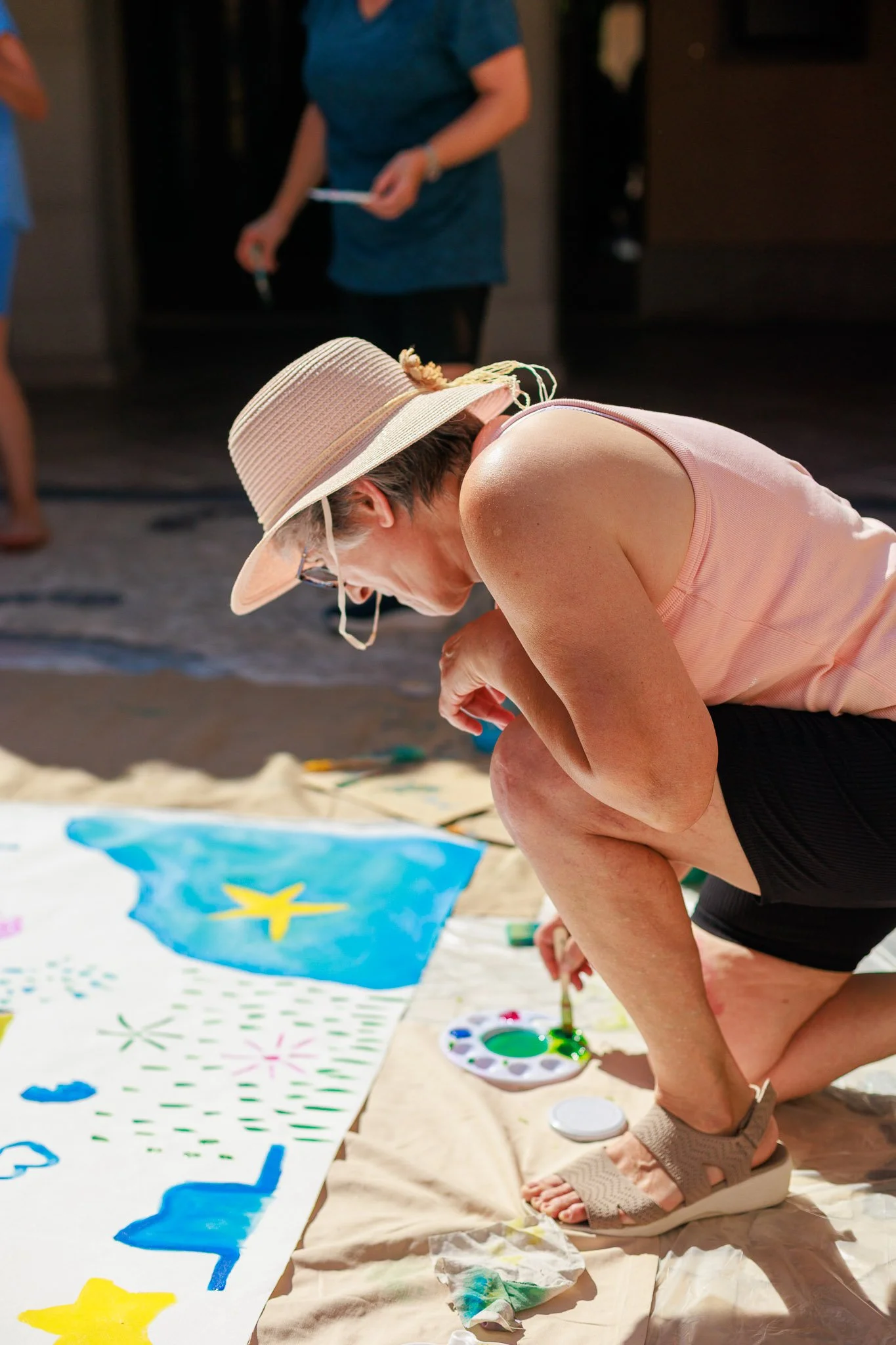 A woman wearing a beige sun hat, glasses, a pink sleeveless top, and black shorts is kneeling on the ground and painting a large outdoor mural on a beige cloth with colorful designs, including a blue star and yellow star, with other painters in the b