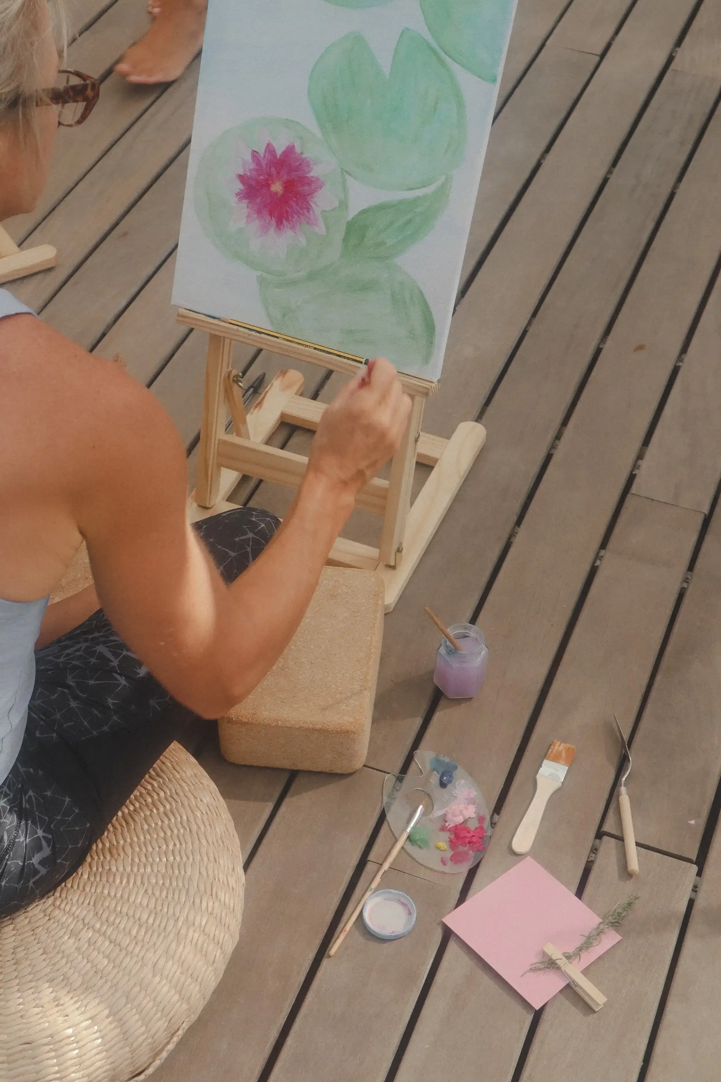 Persona pintando en un caballete con un lienzo que muestra hojas verdes y una flor rosa. Hay pinceles, paleta de pintura, papel rosado y otros materiales de arte sobre un suelo de madera.