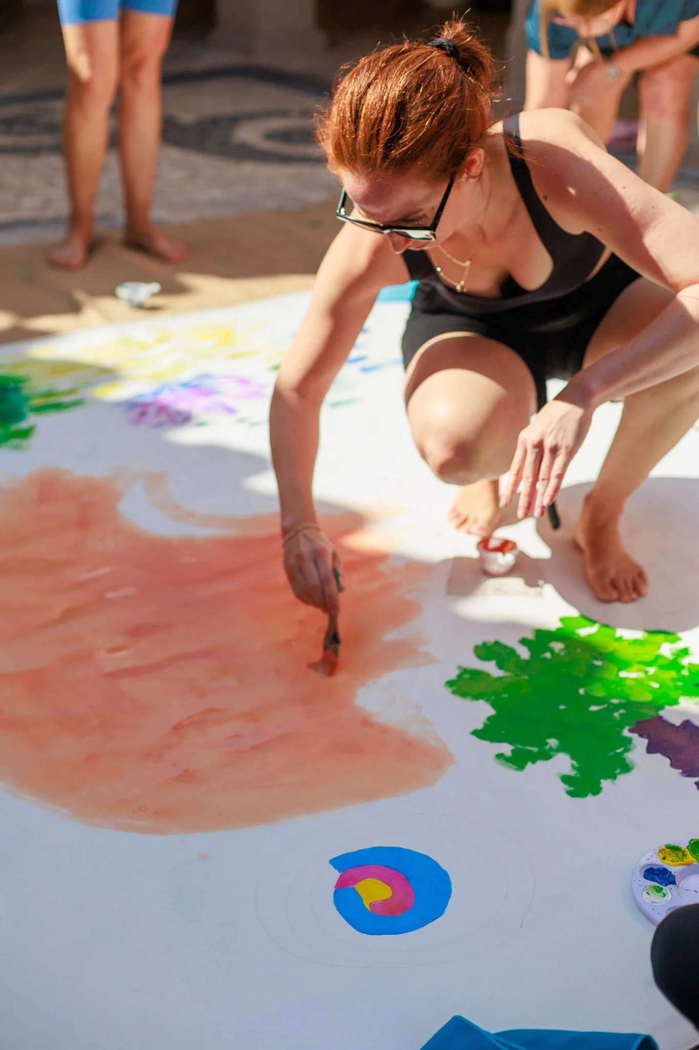A woman crouched on the ground painting on a large white surface, with other people around her. She is using a brush and has various colors of paint nearby.