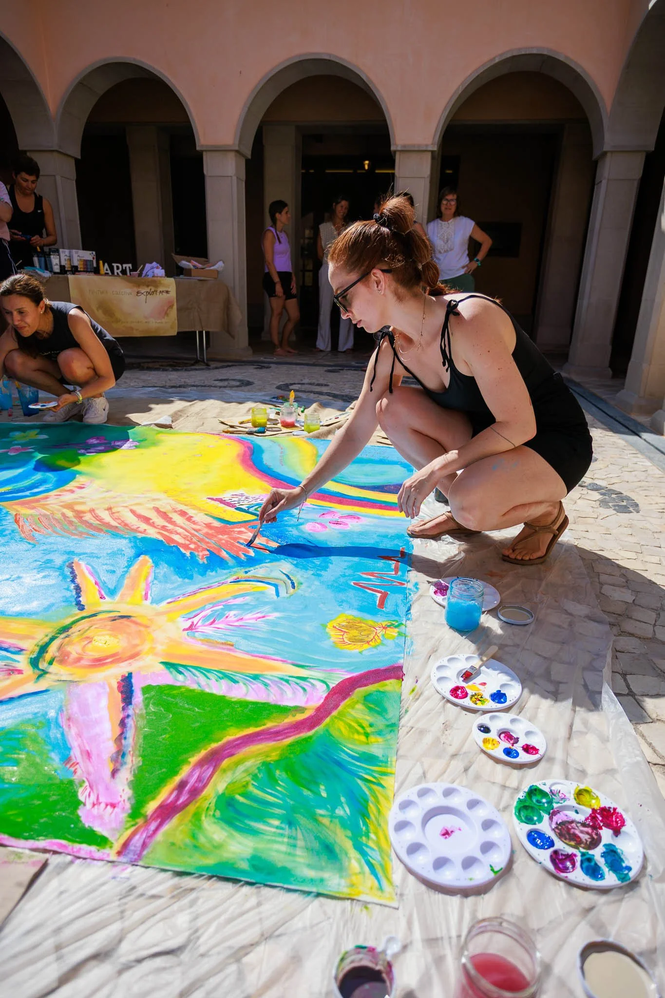 A woman in sunglasses and a black dress crouches on the ground, painting a colorful outdoor mural with bright paints. Several people are in the background near arched buildings.