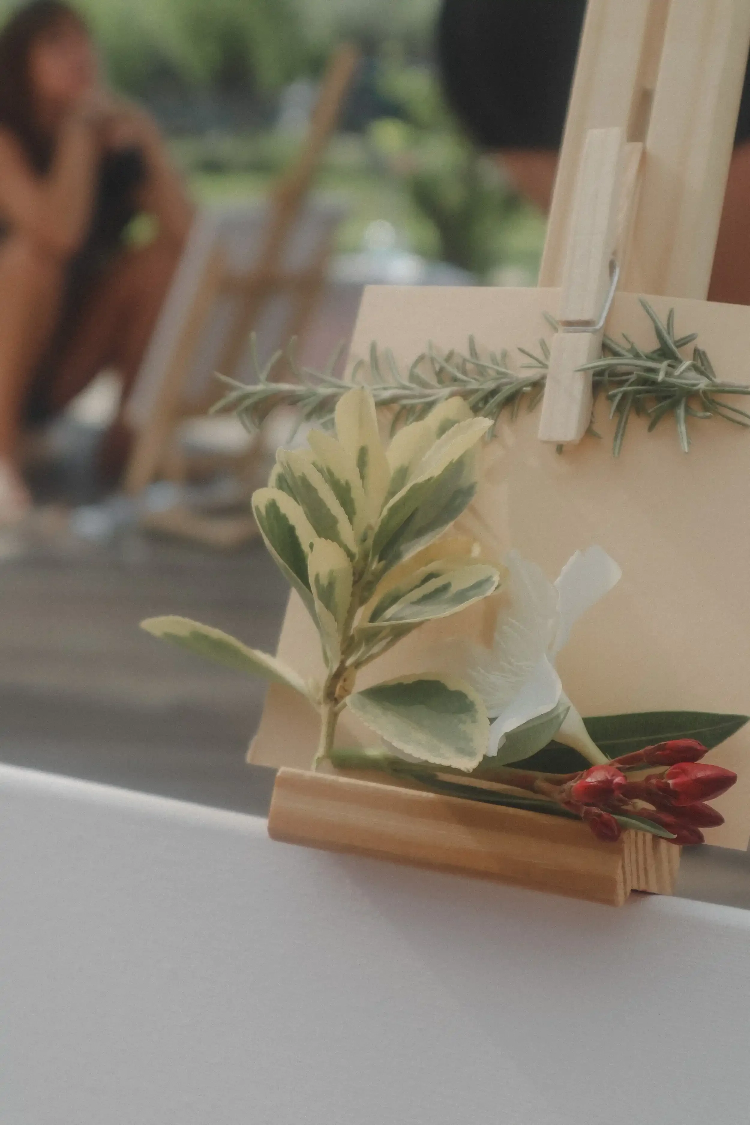 Un caballete pequeño con una tarjeta sostenida por una pinza y decorada con hojas verdes y flores rojas y crema sobre una mesa. Al fondo se ve una figura desenfocada de una persona.