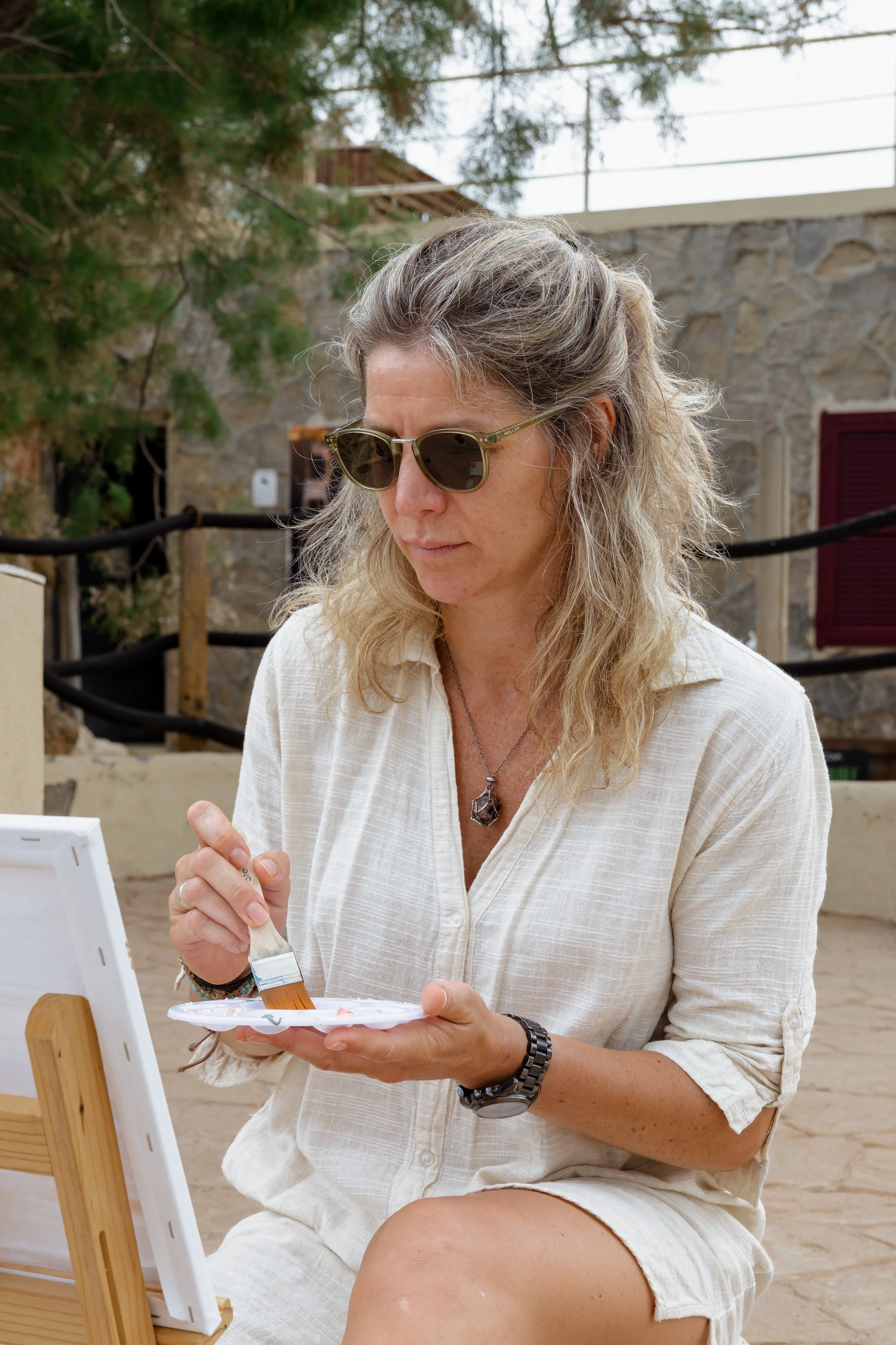 Woman wearing sunglasses painting on a small canvas outdoors.