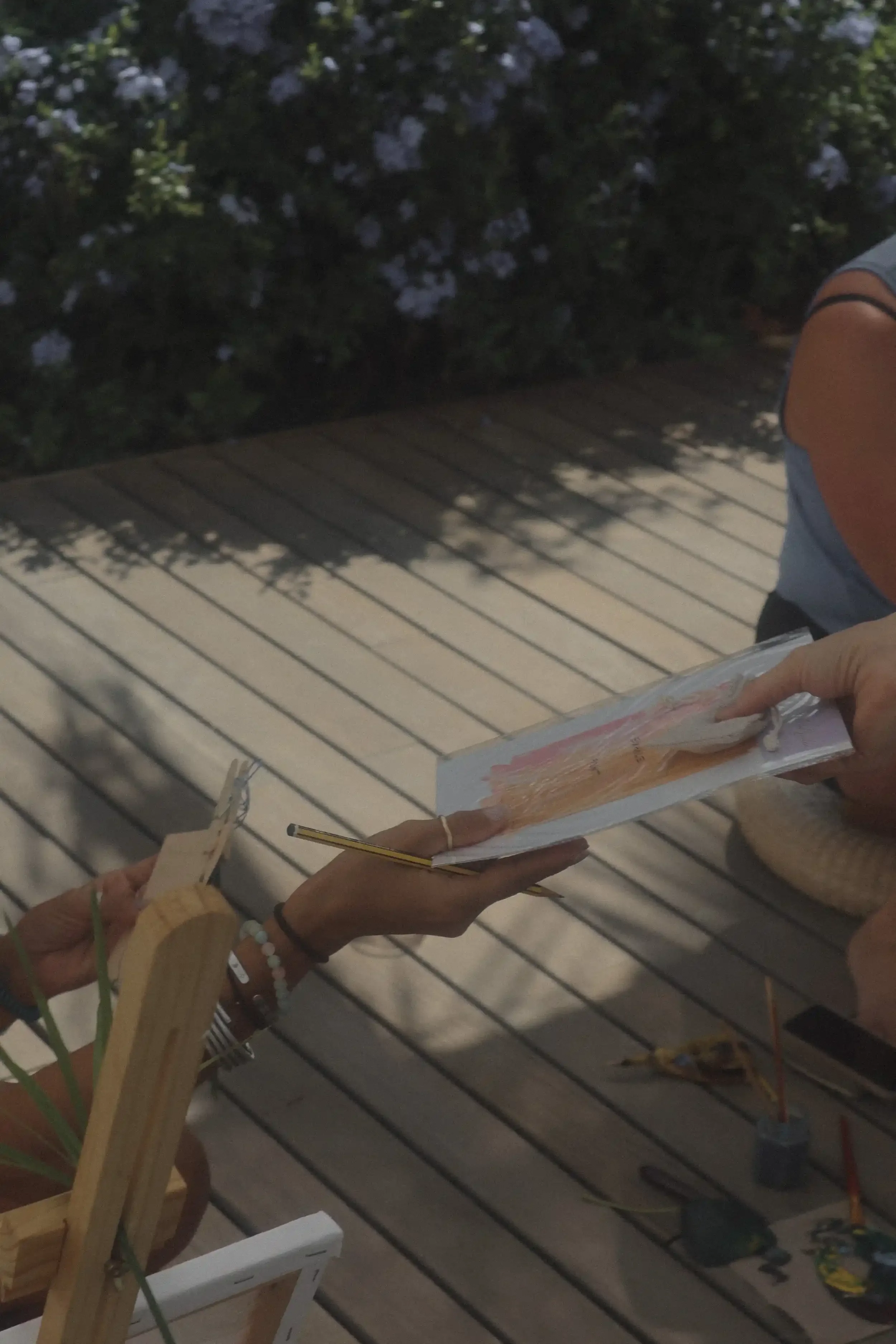 Personas intercambiando una libreta de dibujo en una terraza de madera, rodeadas de materiales de arte.
