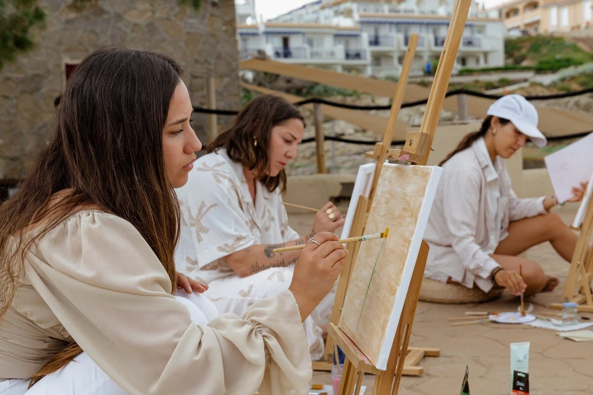 Three women sitting outdoors, painting on canvases placed on easels, with buildings in the background