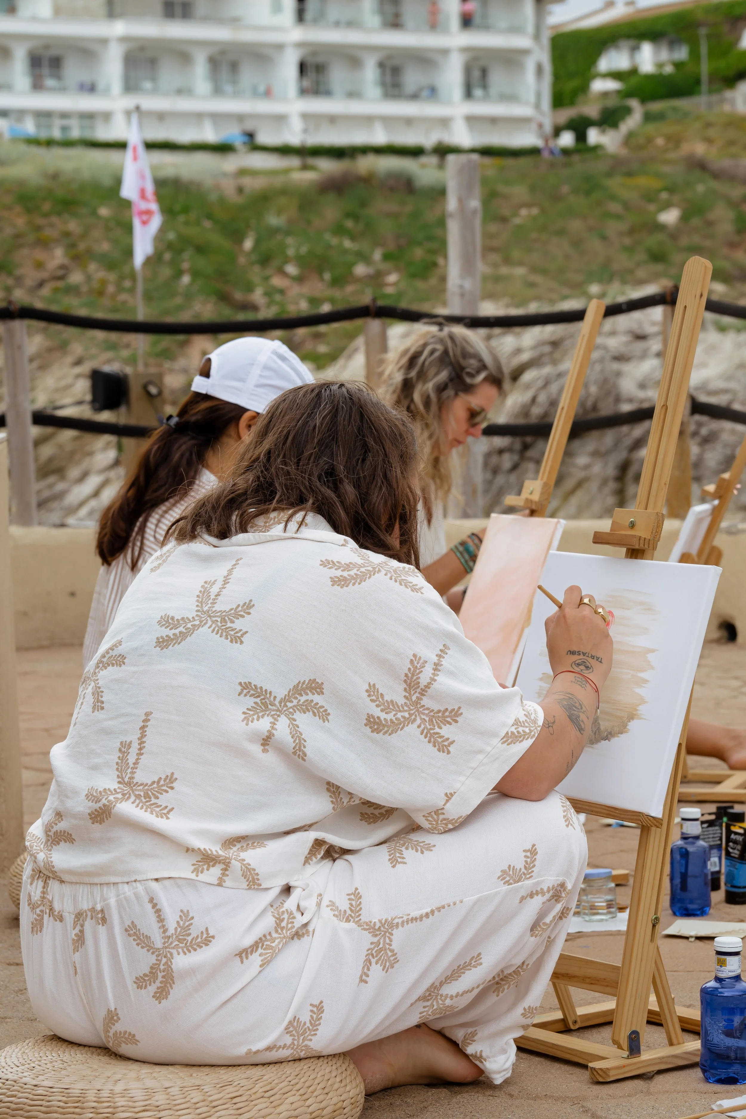 Three women sitting on a sandy surface, painting on canvases set on easels, with bottles of water nearby, outdoors against a rocky background and a large white house with balconies.