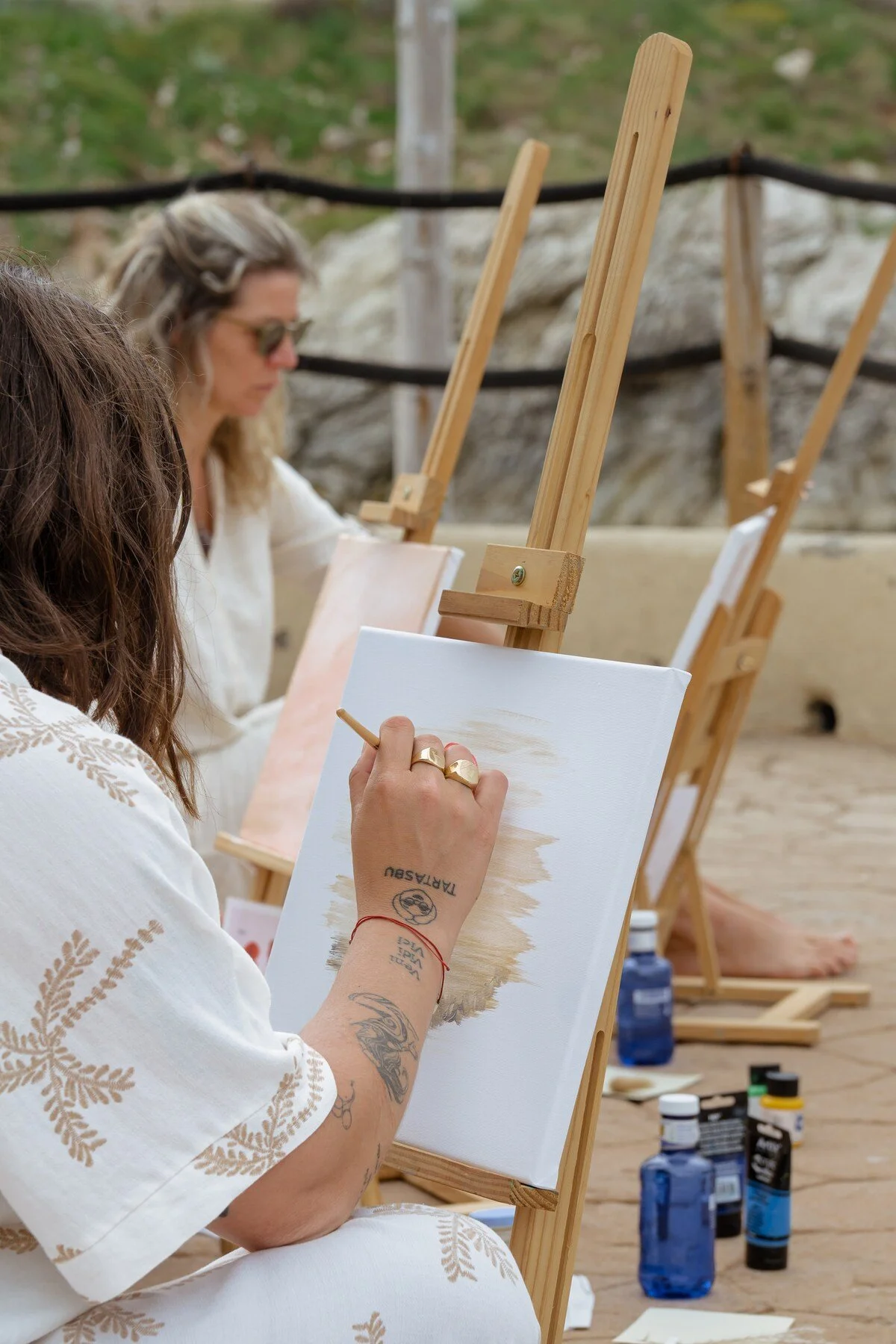 A woman painting on a canvas outdoors, seated on a wooden deck with painting supplies nearby, with another person in the background also painting.