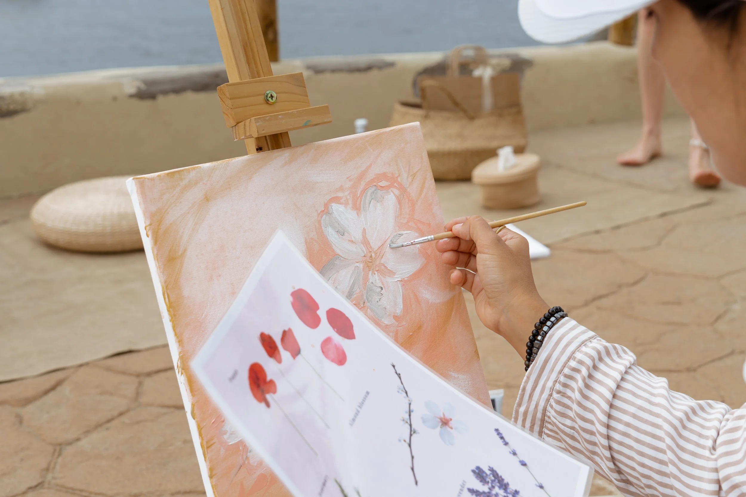 Person painting a white flower on a canvas with a palette of pink, gray, and white colors, at an outdoor art class. The scene includes a wooden easel, a reference sheet, and other art supplies in the background.