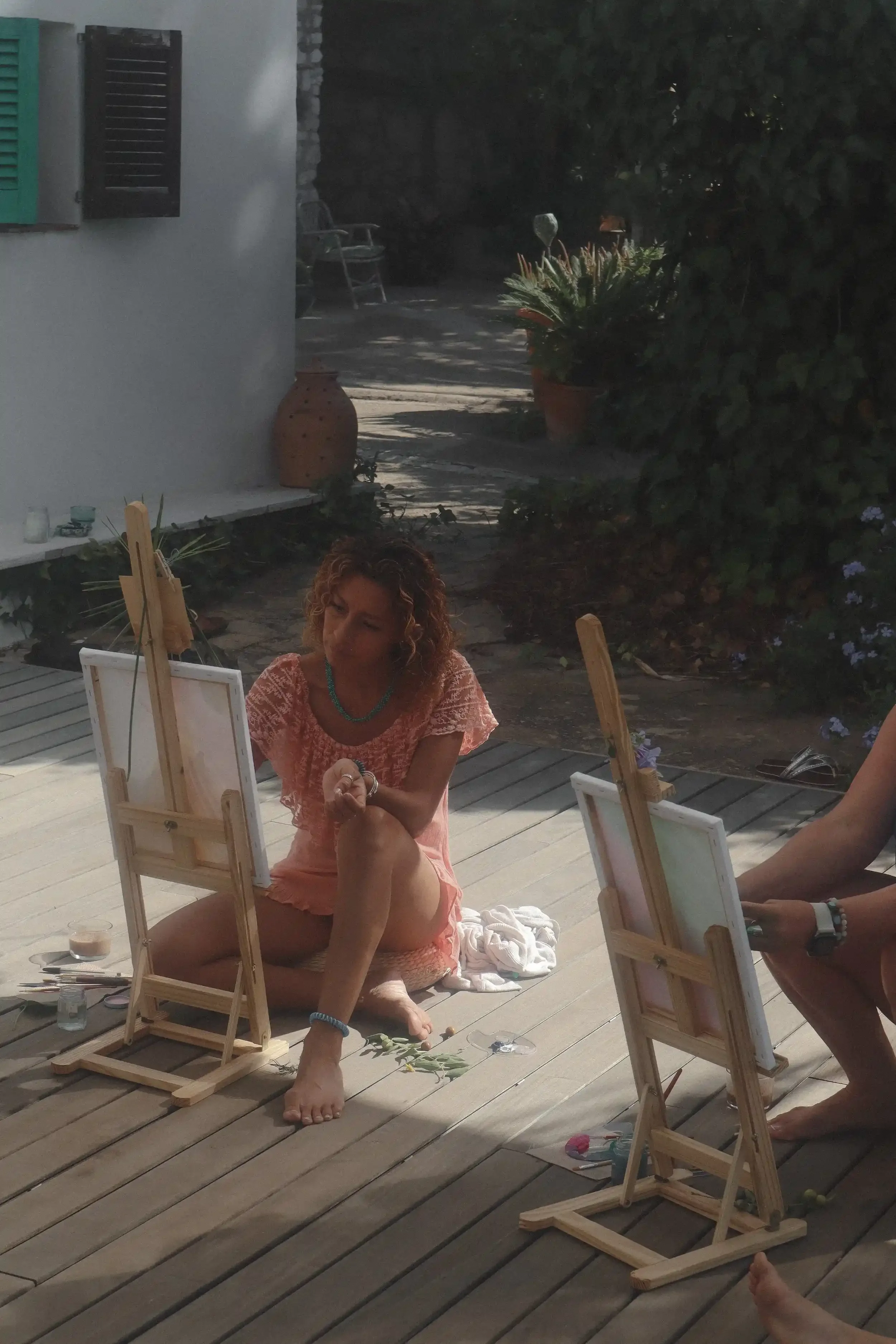 Mujer sentada en el suelo pintando al aire libre en un caballete, rodeada de plantas y luces naturales.