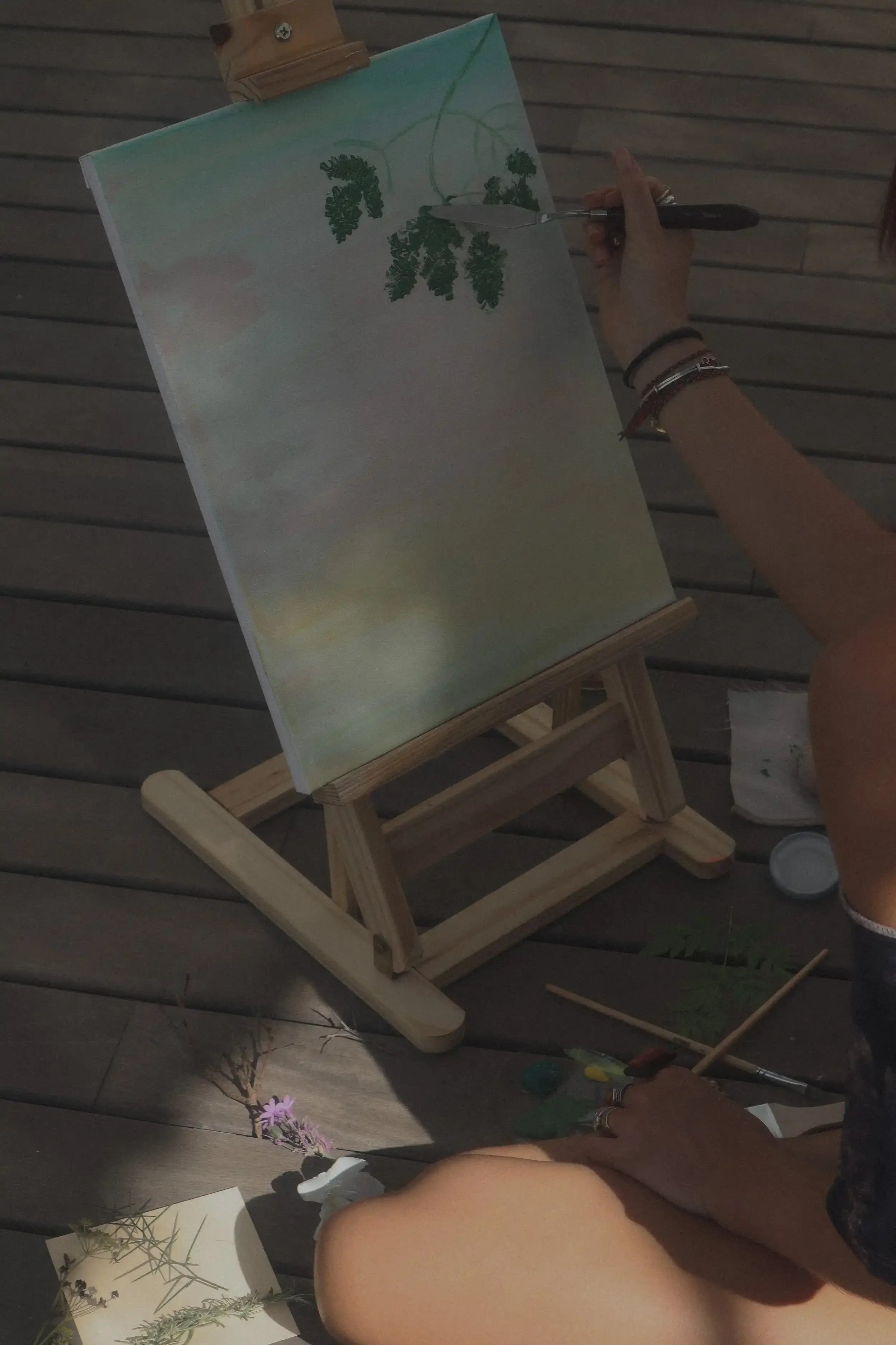 Persona pintando hojas verdes en un lienzo sobre un caballete de madera, en un espacio con piso de madera. Hay utensilios de pintura a su alrededor.