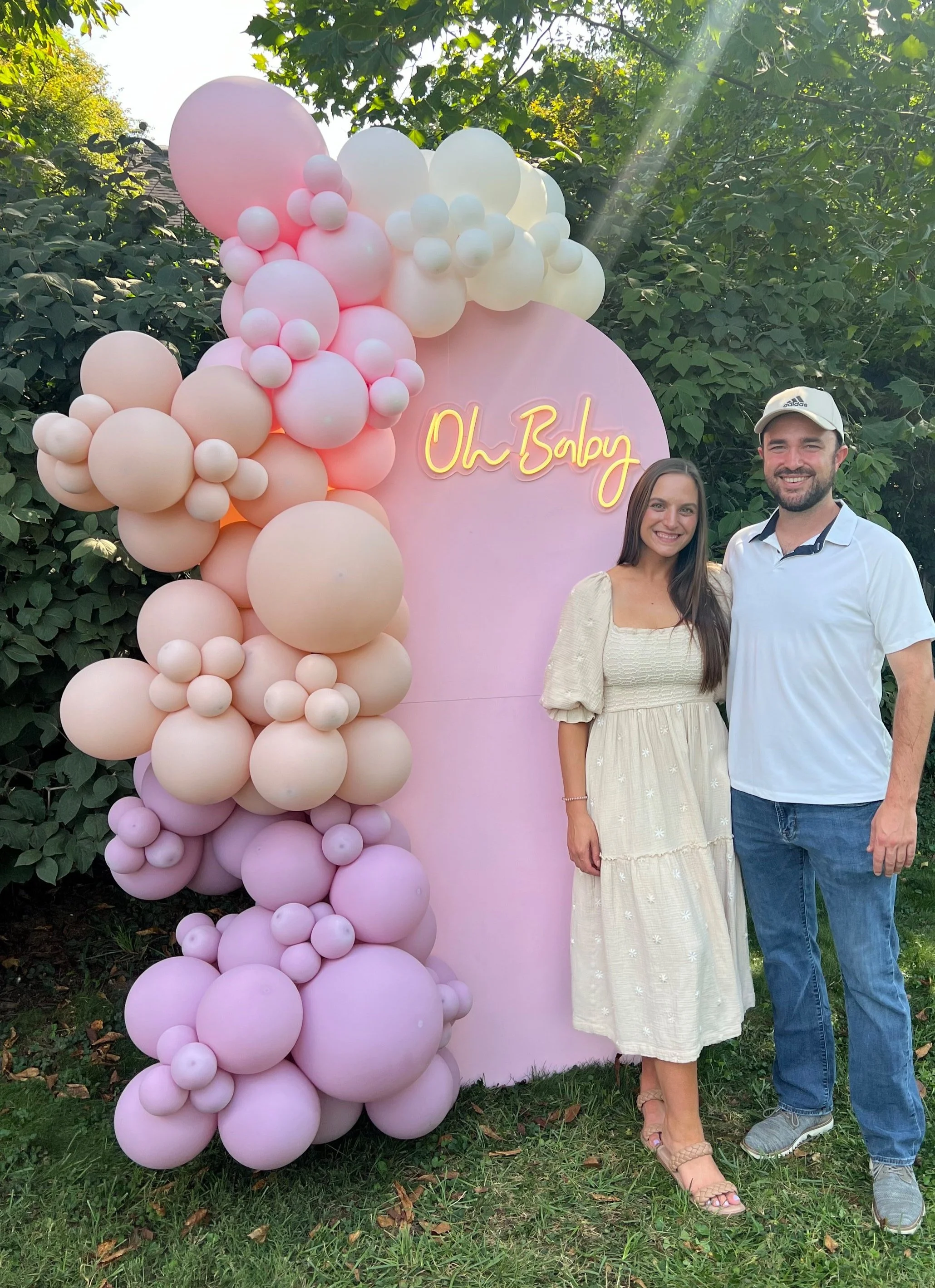 A woman and a man standing together outdoors in front of a pink backdrop decorated with pink and white balloons, with the words 'Oh Baby' in neon lights, surrounded by green trees.