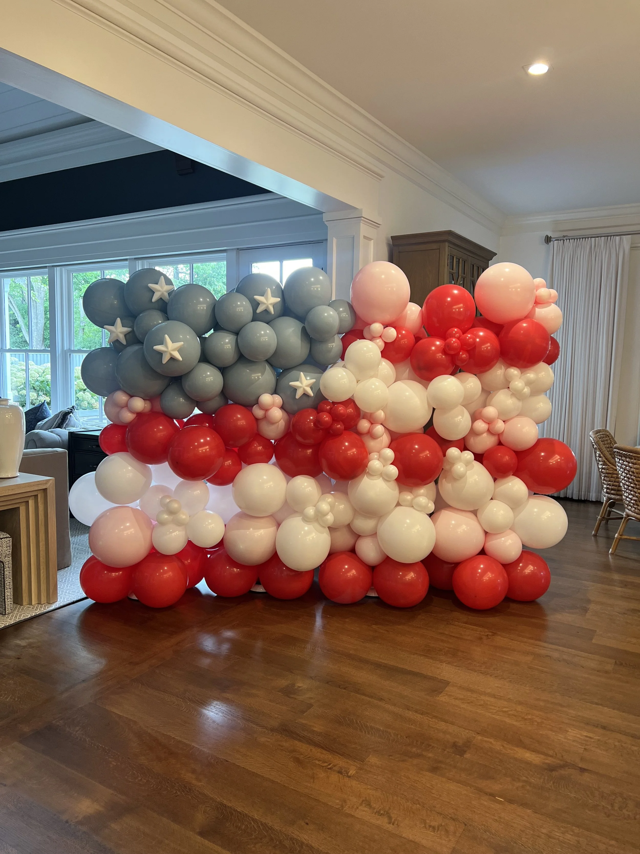 A large decorative balloon wall with gray, red, pink, and white balloons, some with star patterns, arranged in a cluster inside a home.