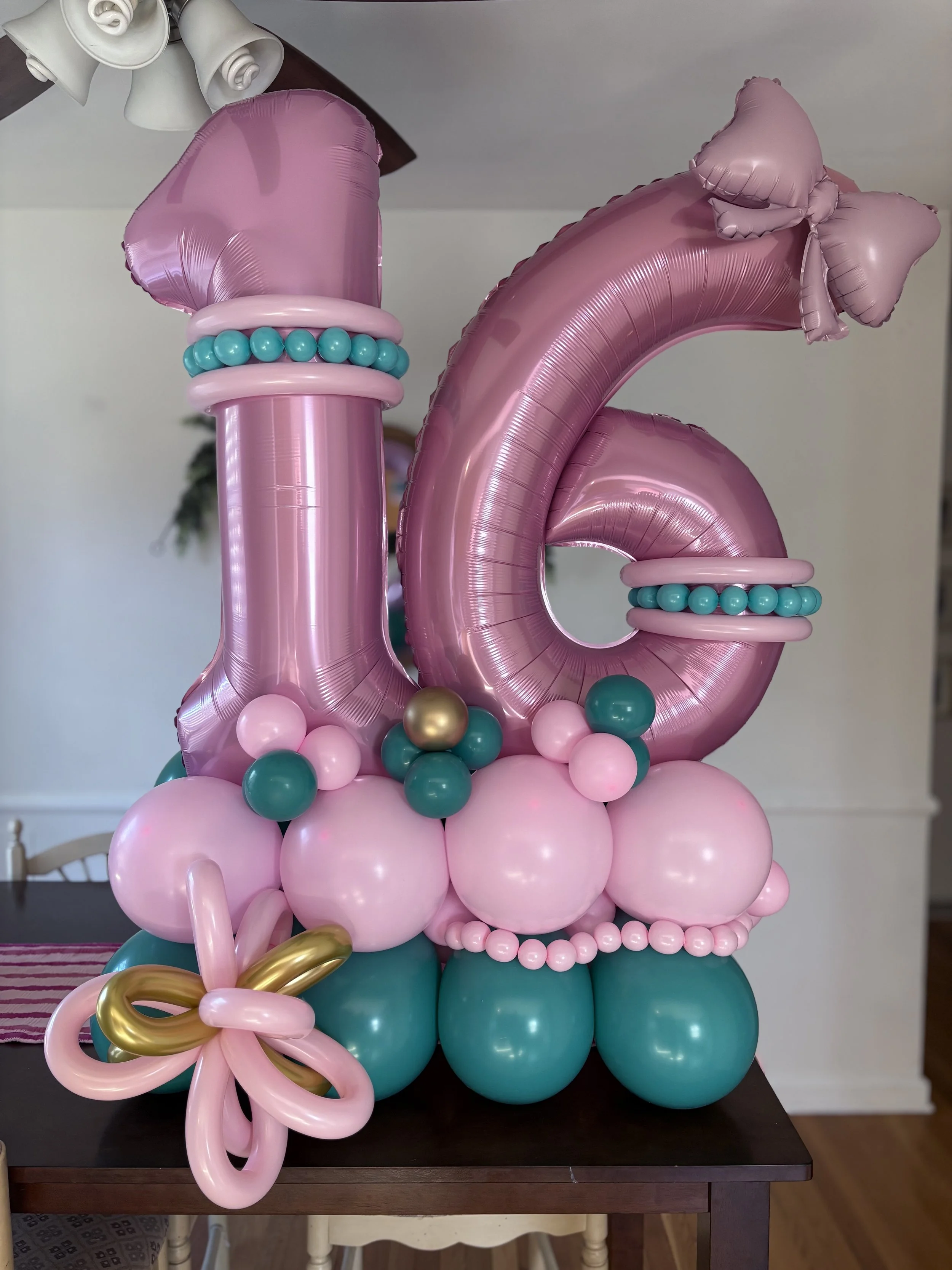 Pink and teal balloon arrangement spelling out the number 16, decorated with bows, beads, and a gold and pink bow, likely for a Sweet 16 birthday celebration.