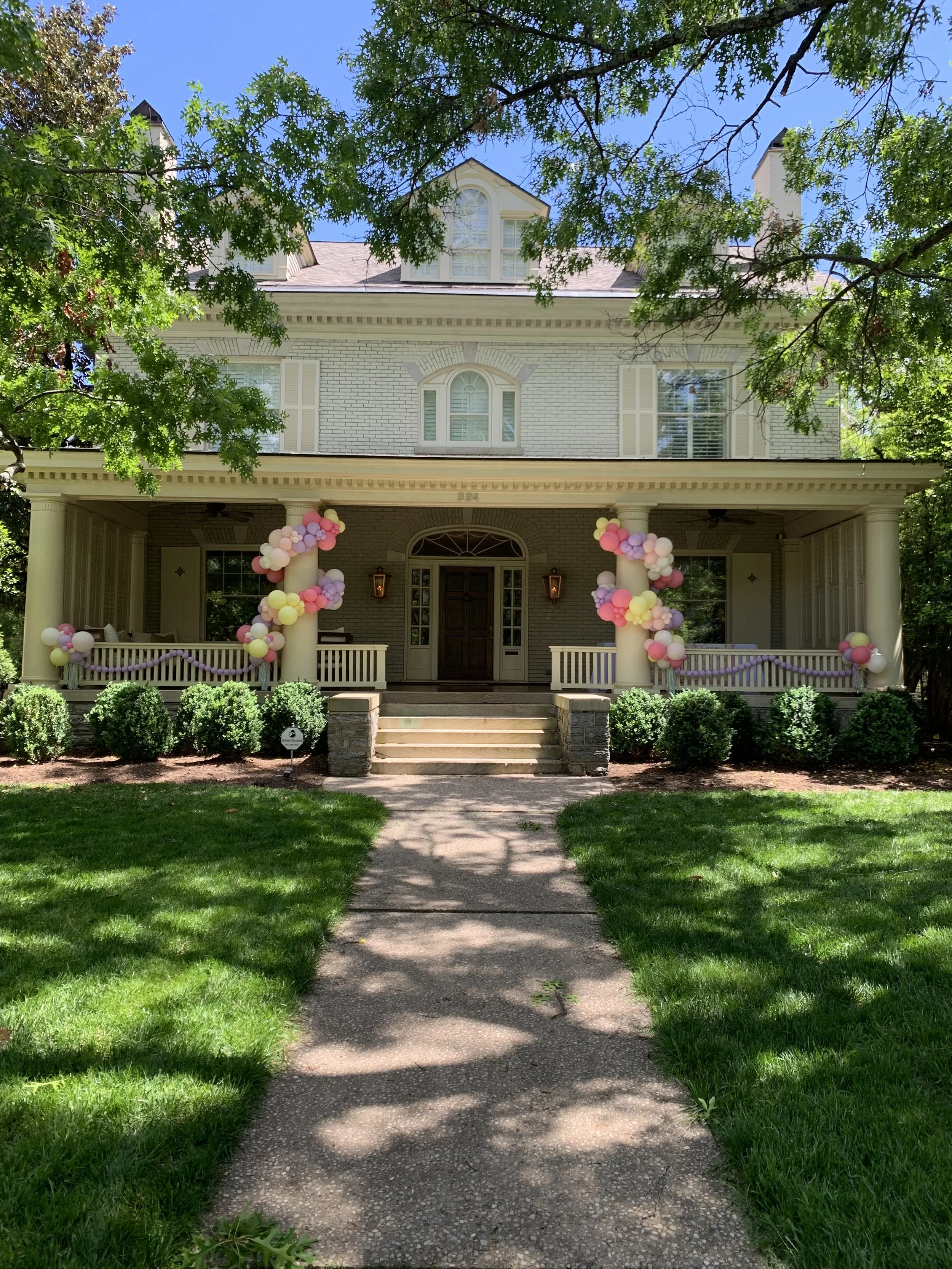 Front view of a large, white, two-story house decorated with pink, purple, and white balloons on columns and railing, with a green lawn, bushes, and a shaded sidewalk leading to the front door.