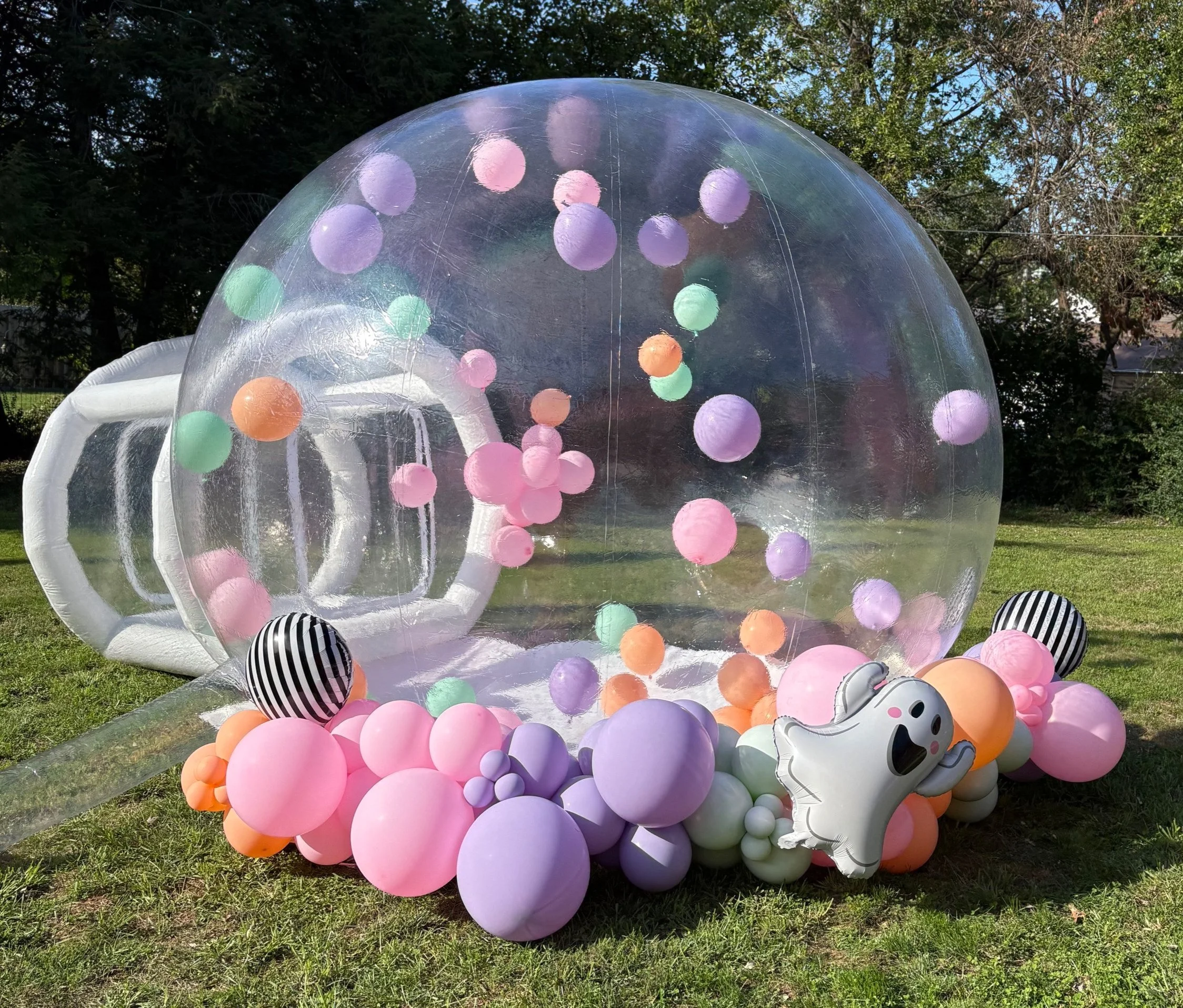 Inflatable clear ball with colorful pastel balloons inside, surrounded by more balloons and ghost-shaped balloon decoration on grass in a backyard.